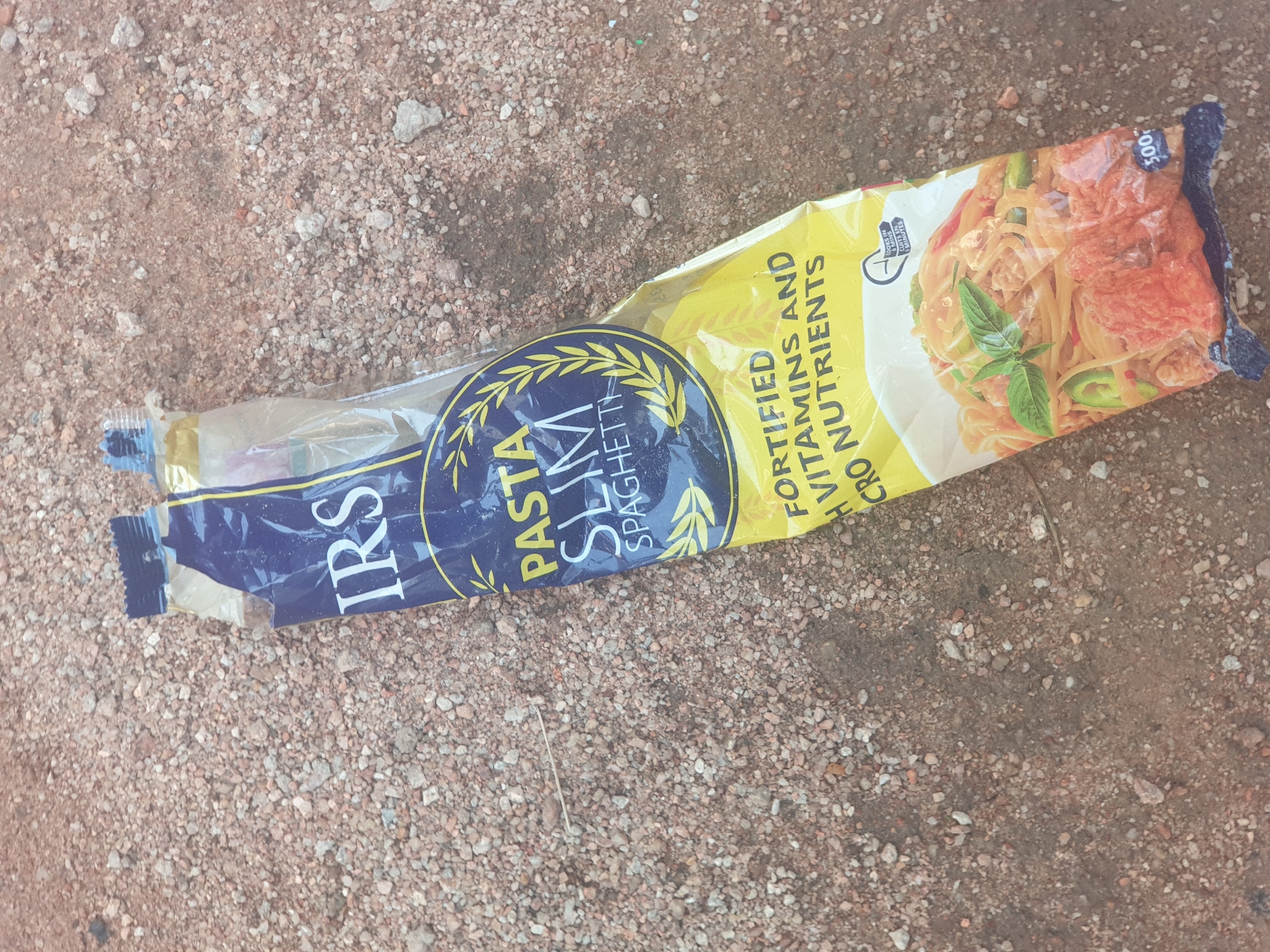 A crumpled bag of IRS brand Pasta Slim Spaghetti lies on a surface of small, reddish-brown stones and dirt. The bag is primarily yellow with blue and white lettering. The text "IRS" is visible in large white letters on a blue background at the top of the bag. Below this, "PASTA SLIM SPAGHETTI" is written in white letters. Further down the bag, "FORTIFIED H VITAMINS AND CRO NUTRIENTS" is printed in white. A picture of cooked spaghetti with vegetables and meat is depicted on the lower portion of the bag. The bag is sealed at the bottom with a black band. The weight "500g" is visible in blue text on the bottom right of the bag. The overall scene is outdoors on a gravelly surface.
