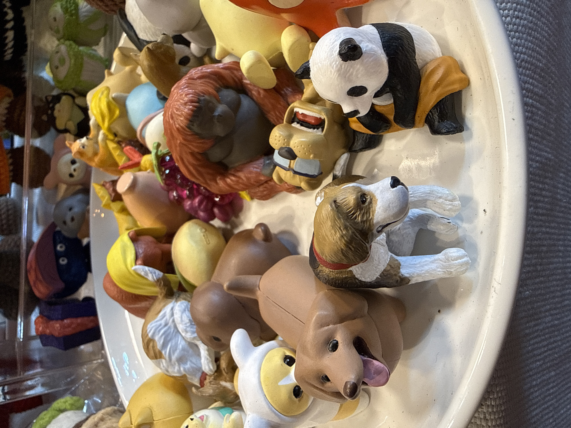 This close-up, slightly overhead shot displays a vibrant collection of small, animal-themed figurines arranged on a white, oval-shaped platter. The scene appears to be indoors, likely in a market stall or souvenir shop in Nha Trang, Vietnam, offering a variety of trinkets.
Prominently featured are several distinct figurines. At the top right, a black and white panda figurine is depicted in a sitting posture with a yellow-orange base. Below it, a tan-colored, dog or lion-like figure with an open mouth revealing teeth and a blue lower jaw rests on its back. Adjacent to this is a large, reddish-brown orangutan figurine, also lying on its back. Further down on the platter, a brown, white, and black beagle-like dog figurine is shown lying, alongside another light brown dog figurine with its pink tongue playfully sticking out. A white and yellow cartoon character and several other colorful figurines (yellow, green, pink, purple) are also visible, some partially obscured.
In the blurred background on the left, additional figurines in various shapes and colors (including green, dark blue/purple, and pinkish hues) are visible within what appears to be clear display shelving. The white platter rests on a textured gray surface, possibly a carpet or fabric. The lighting is uniform, indicating an indoor setting. No discernible text is visible on any of the objects or in the immediate surroundings.