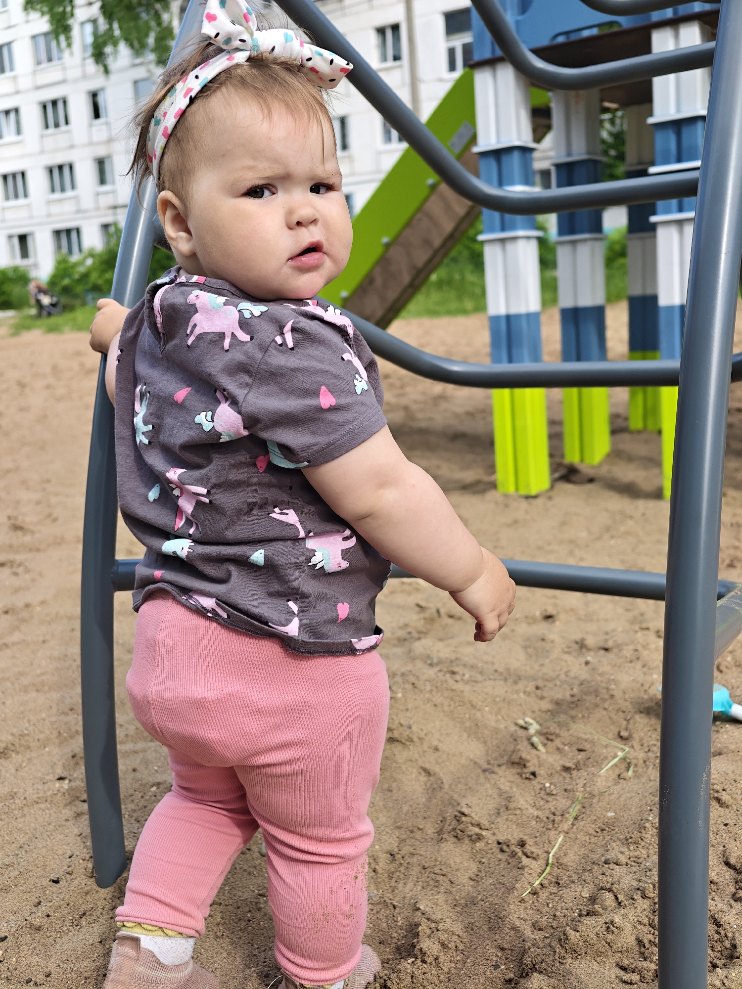 A toddler is depicted on a sandy playground in Chaikovsky, Russia, captured on a bright and sunny day. The child, seen from a slightly angled rear view, looks over their left shoulder directly at the camera with a curious, slightly scowling expression, brows furrowed and lips slightly parted.
The toddler is dressed in a dark grey short-sleeved t-shirt featuring pink and light blue unicorn and heart patterns. They wear pink ribbed leggings and light-colored shoes, with white socks that have a yellow pattern visible. A white headband, decorated with small colorful heart and dot patterns and tied into a bow, adorns their head.
The child is holding onto a dark grey metal bar, which is part of a playground climbing structure. In the background, other brightly colored playground elements are visible, including blue, white, and vibrant green sections that appear to be part of a slide or another climbing apparatus. Beyond the play area, several white multi-story residential buildings with numerous windows can be seen, partially obscured by green trees and foliage. The ground is covered in sand, with occasional blades of grass and small debris. The clear, natural lighting suggests it is daytime, likely morning or afternoon.