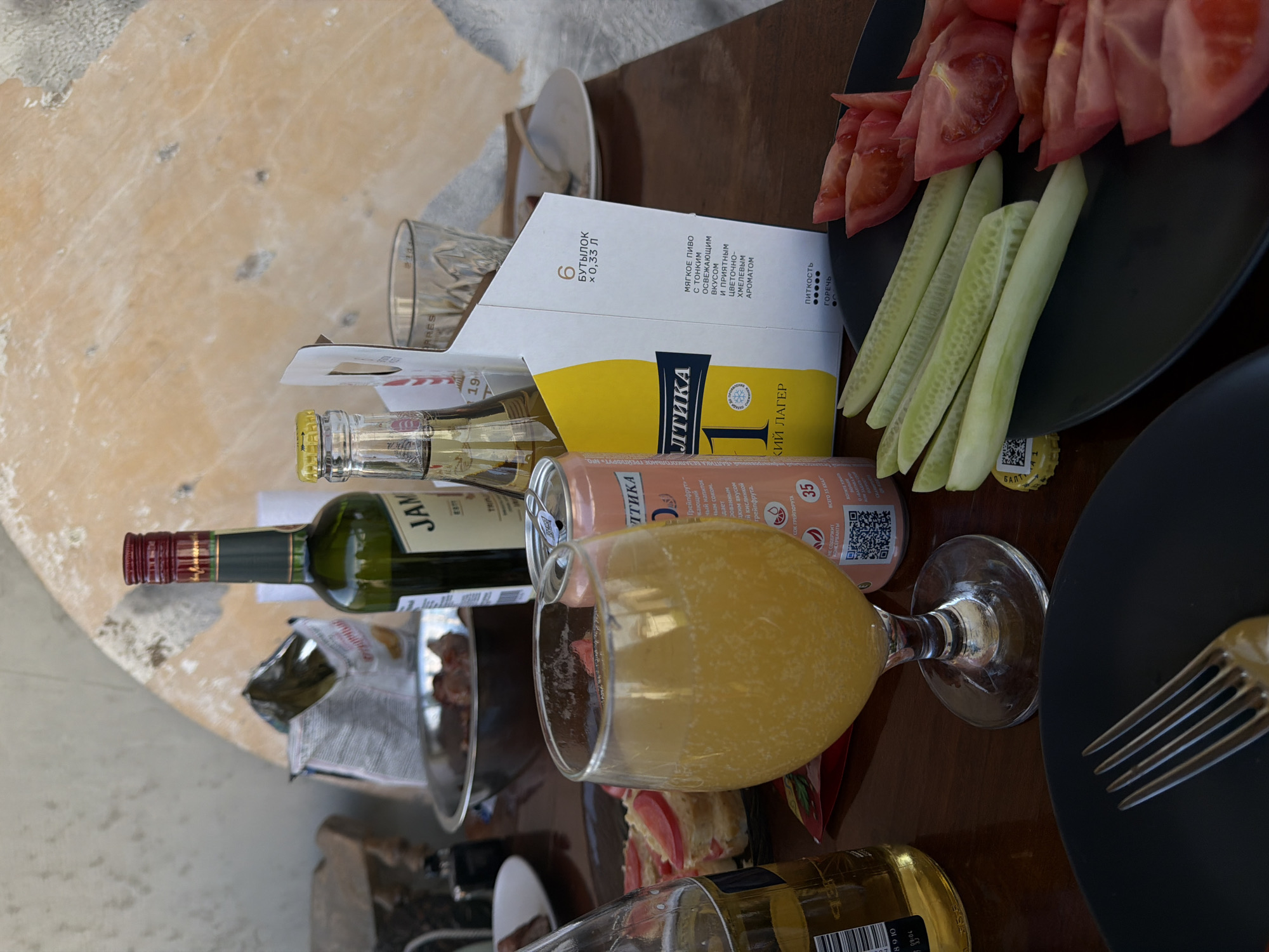 The image captures a casual meal setup on a dark brown table in Chaikovsky, Russia. In the foreground, a stemmed glass contains a bubbly, orange-yellow liquid. To its right, a pink/peach-colored aluminum can displays "БАЛТИКА" (Baltika), "ГРЕЙПФРУТ" (Grapefruit), and a QR code with Russian text. To the left, a partially visible amber glass bottle is present.
Behind these, a green glass bottle with a red screw cap, labeled "JAM," stands prominently. Next to it, a white cardboard carrier specifies "6 БУТЫЛОК x 0,33 Л" (6 bottles x 0.33 L) and features "БАЛТИКА 1 ЛЕГКИЙ ЛАГЕР" (Baltika 1 Light Lager) in Cyrillic, with further descriptive Russian text. An empty, textured clear glass is visible behind the carrier. A discarded yellow bottle cap lies on the table to the bottom right.
A black plate on the right holds sliced green cucumbers and red tomatoes. A silver fork rests on another black plate at the bottom left. In the mid-ground, a silver bowl contains dark, cooked meat. Behind the bowl, an open silver bag, possibly of snacks or chips, is visible. To the left of the bowl, a plate holds small bread slices topped with a white spread and tomato slices.
The background features a light-colored wall, possibly concrete or plaster, with a large, rounded, light brown surface to the right. The scene is illuminated by natural light. No individuals are depicted.