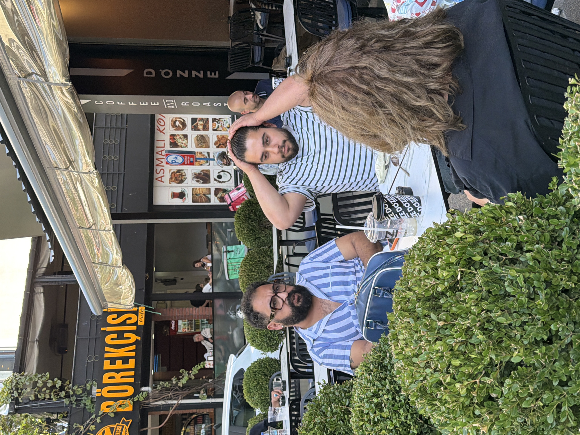The image depicts an outdoor seating area of a cafe in Kadıköy, Turkey. In the foreground, three individuals are seated at a table. A man with dark curly hair, a beard, and glasses, wearing a blue and white striped shirt, is looking to his right. Beside him, another man with dark hair is running his hands through his hair and looking down. A third person, whose back is to the camera, has light brown, wavy hair and is wearing a black shirt. On the table are drinks, cups, and a blue bag. In the background, the cafe's facade is visible, with signage for "BÖREKÇİSİ" and "COFFEE AND ROAST." Other patrons are seated at tables, and a car is parked on the street. Lush green bushes surround the seating area.