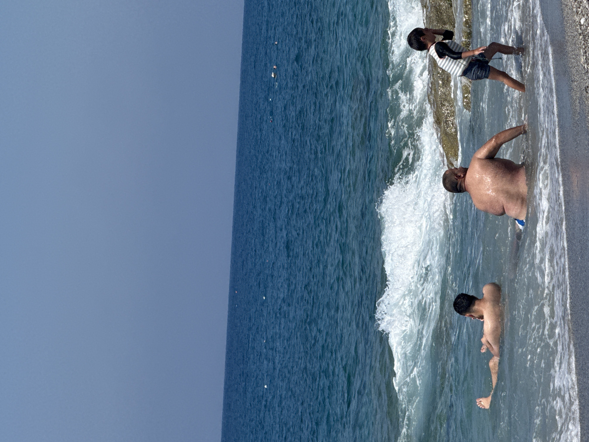 The image depicts a sunny day at the beach in Alanya, Turkey, with people enjoying the sea. In the foreground, a young boy wearing a striped rash guard and dark shorts is walking towards the right, shielding his eyes from the sun. To his left, a man with a large build is seated in the shallow water, facing away from the camera. Further to the left, another man is lying in the water, with his back to the camera. The ocean is a deep blue, with waves crashing on the shore. Rocky outcrops are visible in the water behind the people. The sky is a clear, pale blue.