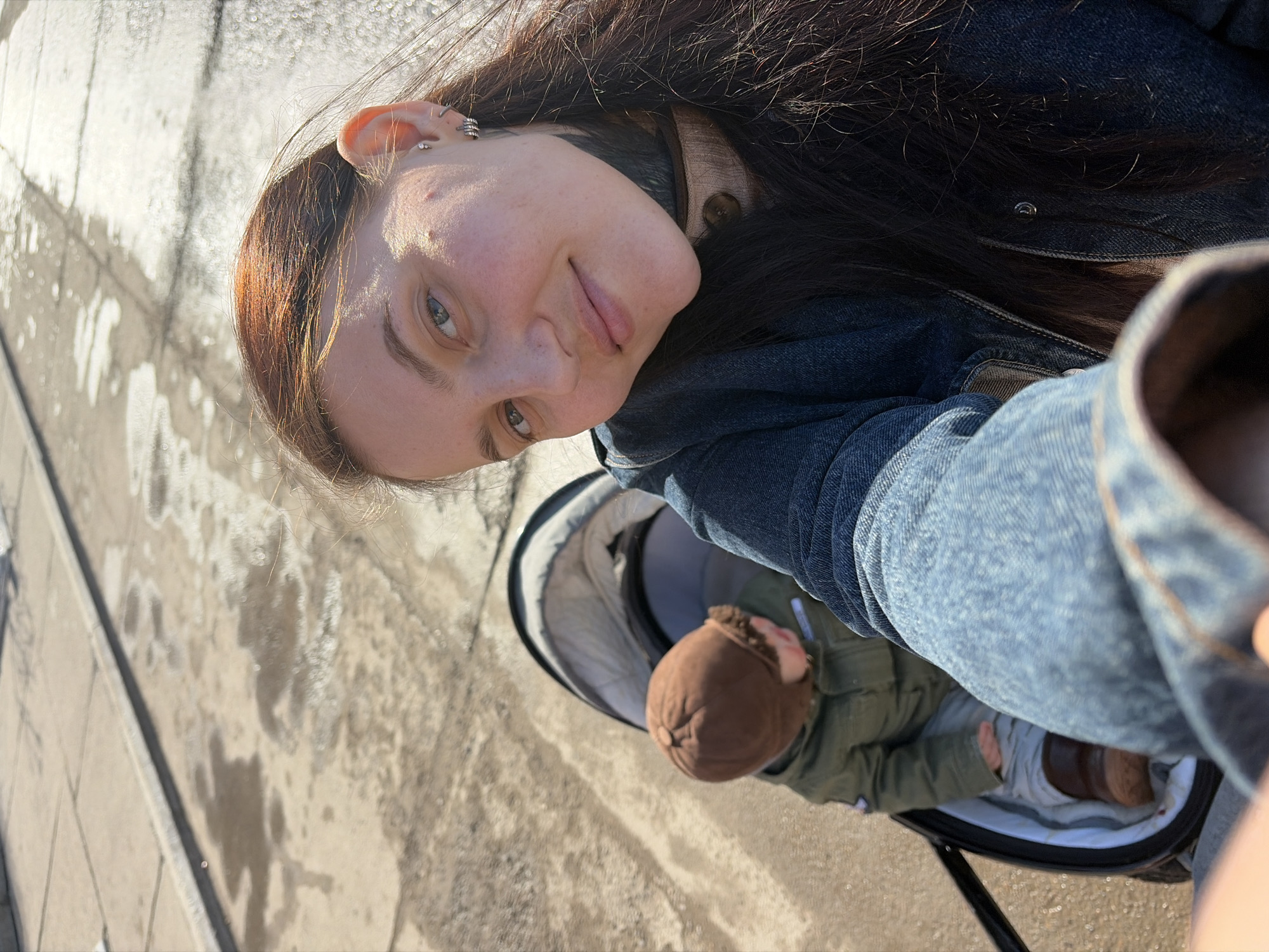 An adult female is captured in a selfie with a child in a stroller, outdoors in the city of Chaikovsky, Russia. The woman, foregrounded and facing the camera, has long dark hair with reddish highlights, light-colored eyes, and multiple piercings in her visible left ear, including studs and hoops. She wears a dark denim jacket over a light-collared garment and a dark neck accessory. Her expression is a slight smile. To her right and slightly behind, a young child is seated in a dark-colored stroller with a light grey interior. The child wears a brown aviator-style cap, a green padded jacket, light-colored pants, and brown boots, facing away from the camera and looking downwards. The ground surface is paved, likely asphalt or concrete, exhibiting patches of standing water and strong reflections from bright sunlight, indicating a wet condition, possibly due to melted snow or recent rain. The scene is brightly lit by direct sunlight.