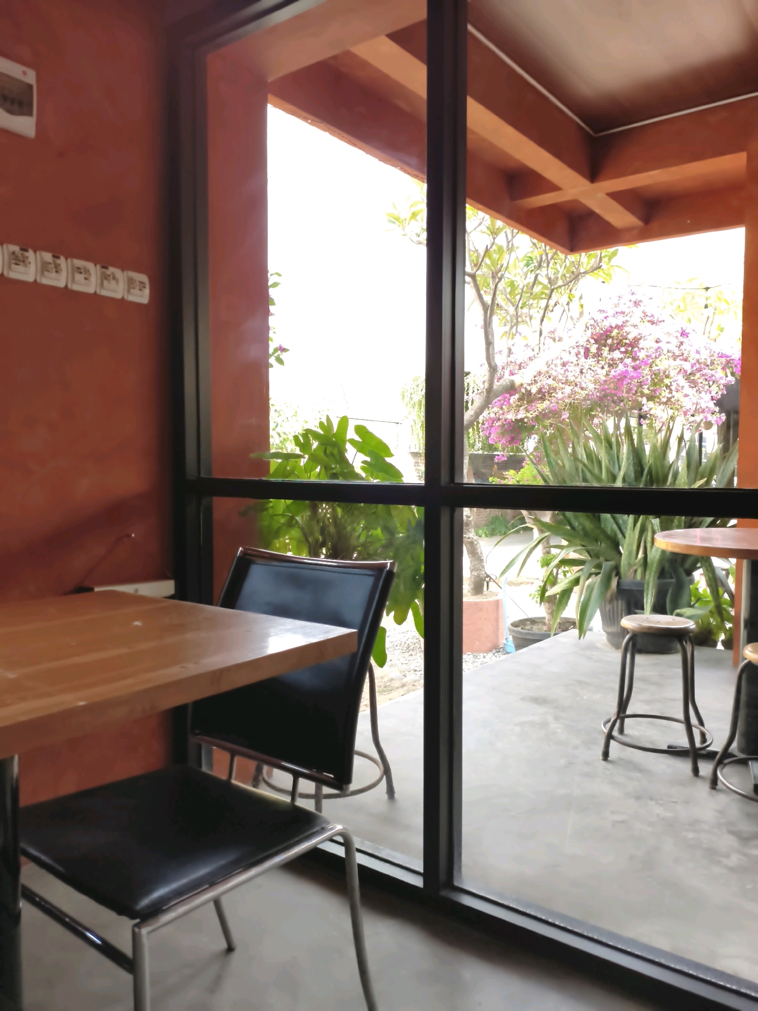 The image shows an interior view of a cafe or restaurant looking out through large glass windows to an outdoor seating area with plants. In the foreground, a wooden table with a dark upholstered chair is positioned to the left. Through the window, the outdoor area features concrete flooring, various potted plants including large snake plants and a flowering tree with pink blossoms, and several tall stools around round tables. The architecture visible suggests a warm climate with exposed wooden beams on the ceiling. The scene appears to be located in Ronowijayan, Indonesia, indicated by context not visible in the image itself. The lighting suggests it is daytime.