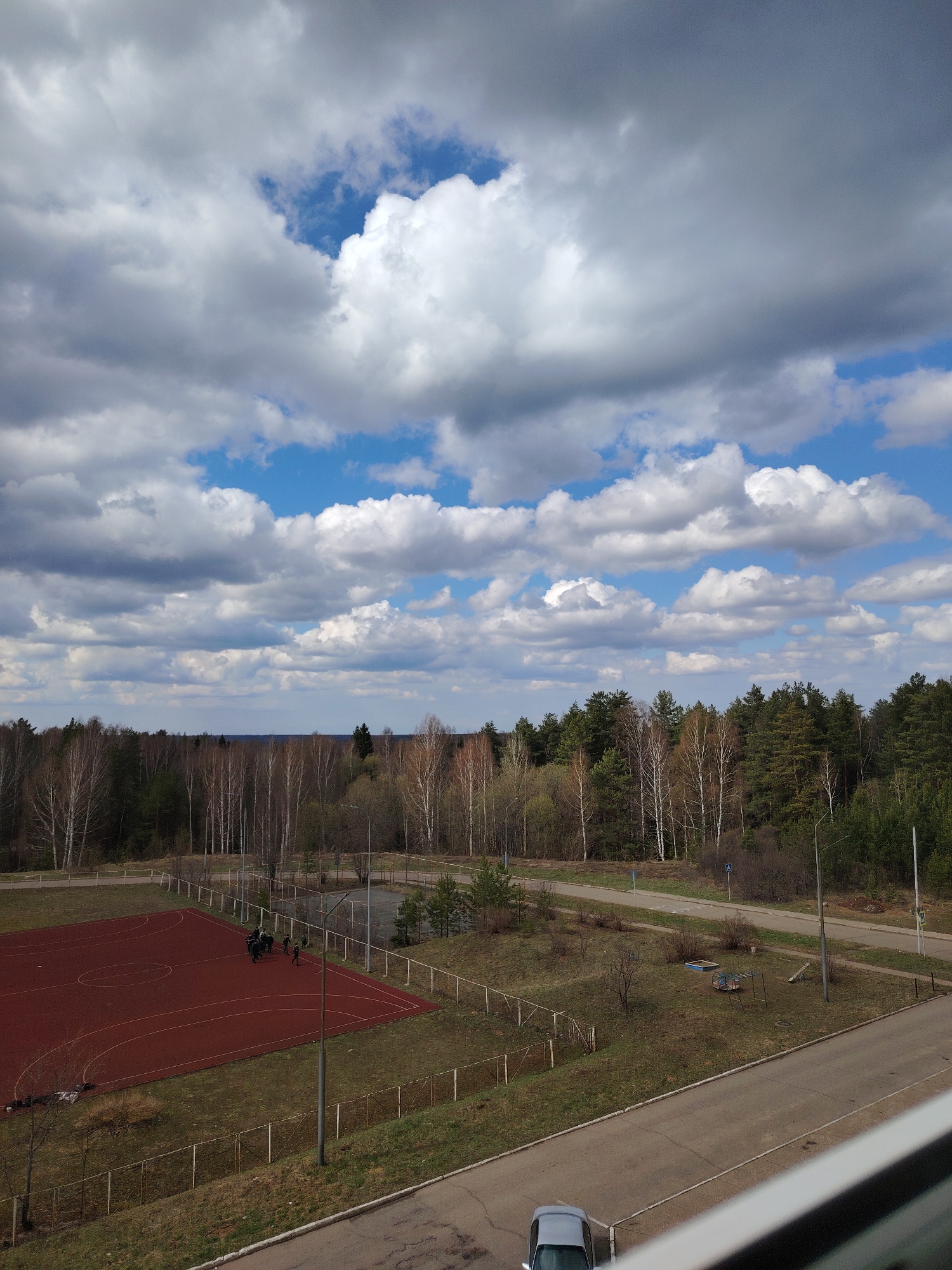 The image displays an elevated view of an outdoor landscape in Марковский, Russia, seen from a high vantage point, possibly a building window. The upper half of the frame is dominated by a dynamic sky filled with large, fluffy cumulus clouds in various shades of white and grey, interspersed with clear patches of vibrant blue sky, indicating partly cloudy weather on a bright day.
Below the horizon, a dense forest stretches across the mid-ground, composed of both deciduous trees, many of which are still bare or show only nascent green leaves, and darker evergreen conifers, suggesting the season is early spring. In the foreground, a vibrant red sports court, marked with white lines for what appears to be basketball or futsal, is a prominent feature. A small group of approximately six to eight people, dressed in dark clothing, are gathered casually on this court.
Surrounding the court and winding through the scene are asphalt roads and unkempt grassy areas, delineated by fences. A small, simple playground featuring a sandbox and a seesaw or similar equipment is visible near the roads. Utility poles stand along the roadside. At the very bottom of the frame, the front portion of a light-colored car is partially visible, parked on a road. The scene portrays a quiet, community area bordering a natural forest, under pleasant daytime lighting. No visible text or signage is legible within the image.