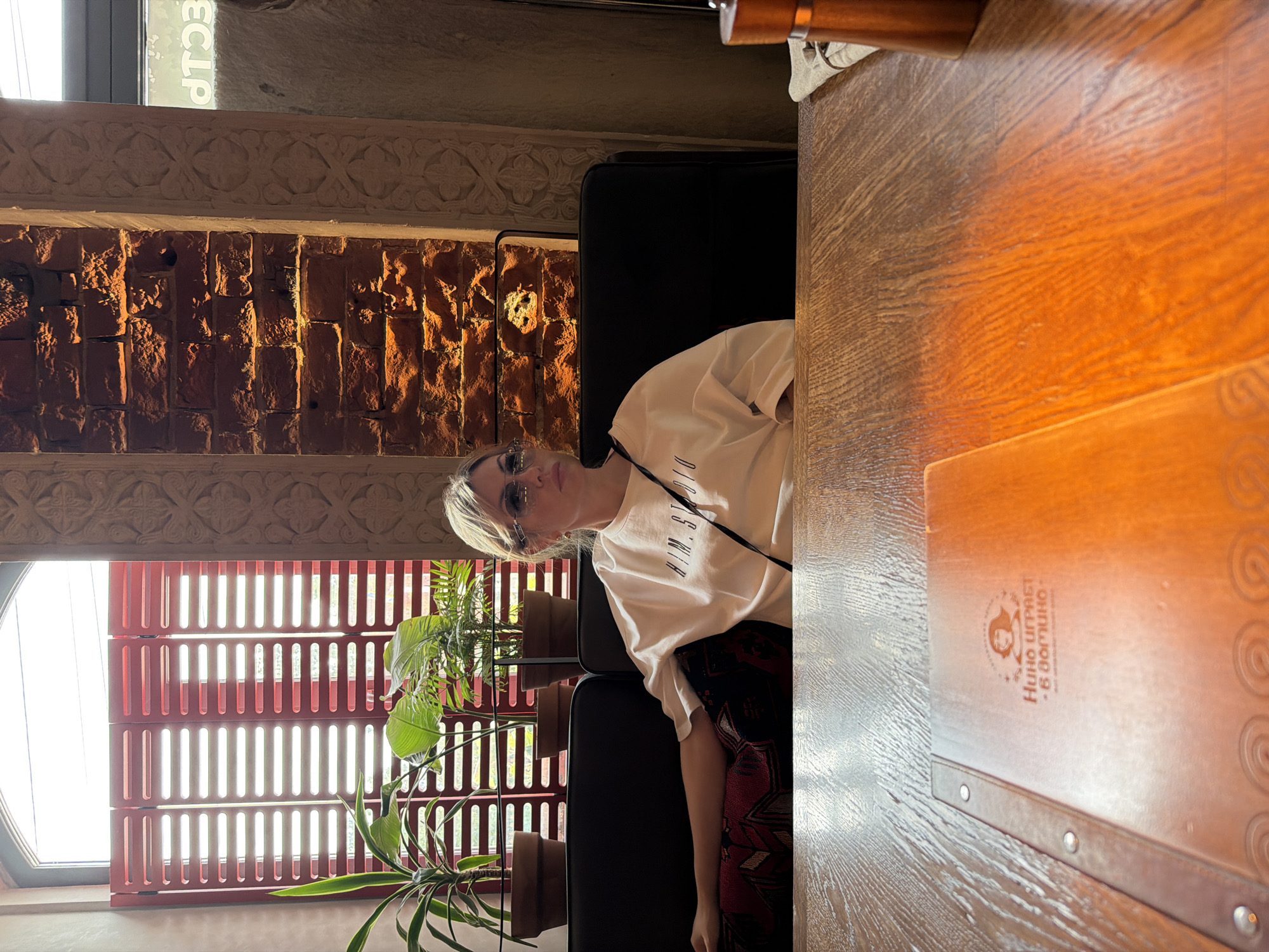 An eye-level, full shot captures a woman sitting at a table in a restaurant. She is wearing sunglasses and a white t-shirt, and her arm is resting on the back of the booth. The woman is looking to her right, away from the camera. In the foreground, a wooden table holds a menu with Russian text and an image of a woman's profile. Behind the woman, a black booth lines the wall, which is adorned with exposed brick and decorative molding. A window with red blinds is visible to the left, and plants are placed on a windowsill. The lighting is warm, with shadows cast by the brickwork. The environment suggests a cozy, interior dining setting.