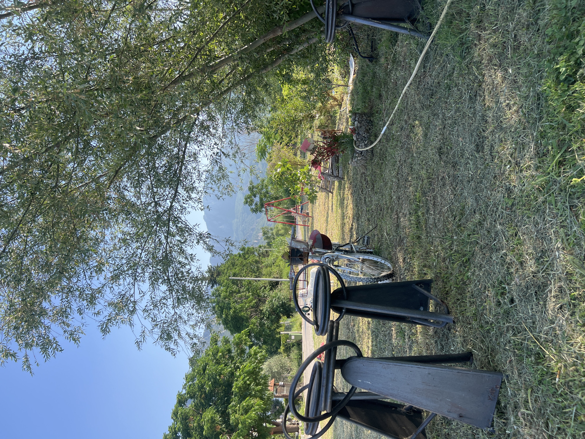 An outdoor scene in Šušanj, Montenegro, features a grassy area with several elements. In the foreground, two metal bar stools with circular tops and angled legs are positioned on the grass. To their right, a white bicycle with a red basket on the handlebars and a red bag on the rear rack is parked. Beyond the bicycle, a red swing set with two swings stands on the lawn. Further back, there are lush green trees and shrubs, with a hazy mountain range visible in the distance under a clear blue sky. A yellow hose is visible on the ground in the mid-ground. The overall impression is a relaxed, outdoor setting.
