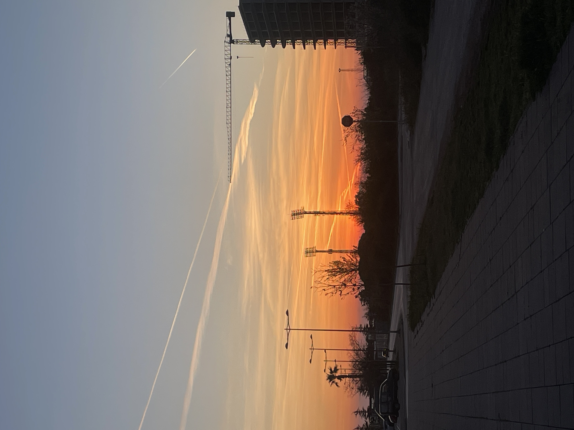The image shows a street scene at sunset in Bar, Montenegro. The sky is a gradient of orange, yellow, and blue, with multiple airplane contrails visible. A construction crane and an unfinished concrete building are present on the left. Stadium lighting is visible further down the street. Streetlights line the road, and silhouettes of trees and buildings are visible against the sunset. A paved sidewalk and curb are on the right, with grass and bushes. The light suggests the photo was taken in an urban, likely residential area of Bar. A row of parked vehicles is visible.