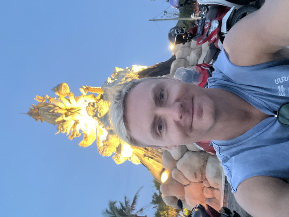 The scene depicts an outdoor environment in Kerobokan Kelod, Indonesia, captured during twilight or early evening. In the foreground, a Caucasian male, appearing to be in his 30s to 40s, with short blond hair, is centered, looking directly at the camera with a slight smile. He is wearing a light blue sleeveless t-shirt, with dark sunglasses hooked onto the collar.
Immediately behind the individual, and forming the central background, is a large, multi-tiered monument, intensely illuminated by warm yellow light. The monument features intricate, gold-toned sculptural elements, including figures with multiple arms and ornate spires at its apex, resting on a base composed of large, rounded, light-colored stones or sculpted elements.
To the far left, a palm tree is visible. To the right of the foreground subject, several motorcycles are parked or idling. A red motorcycle with a clear windshield is partially visible behind the man's left shoulder. Further to the right, another red motorcycle is visible, featuring a black helmet with a skull design hanging from its handlebars, and a partial Indonesian license plate reading "DK 30XX BGS". A white motorcycle is partially visible beyond this. In the deeper background, faint, blurred figures of other individuals are discernible, and a street light emits a glow. The sky above is clear and light blue, transitioning to a darker hue.