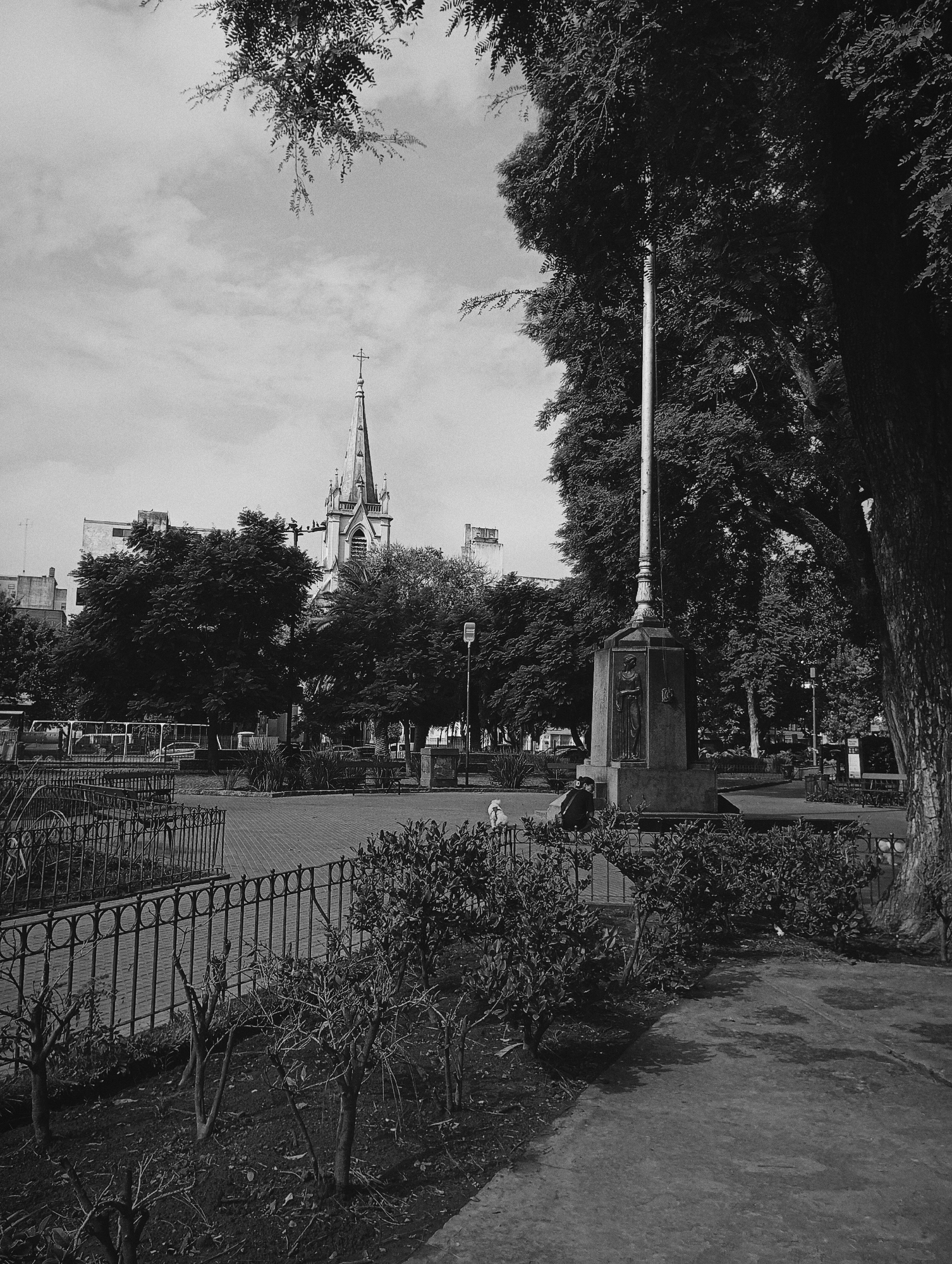 This is a black and white photograph of Plaza Mitre in Buenos Aires, Argentina. In the background, the spire of the Parroquia Inmaculada Concepción de Belgrano is visible. In the foreground, a metal fence encloses a planting bed. To the right, a tall monument features a figure and a vertical pole. A person sits near the base of the monument next to a dog. The sky is overcast. Trees provide shade to the plaza.