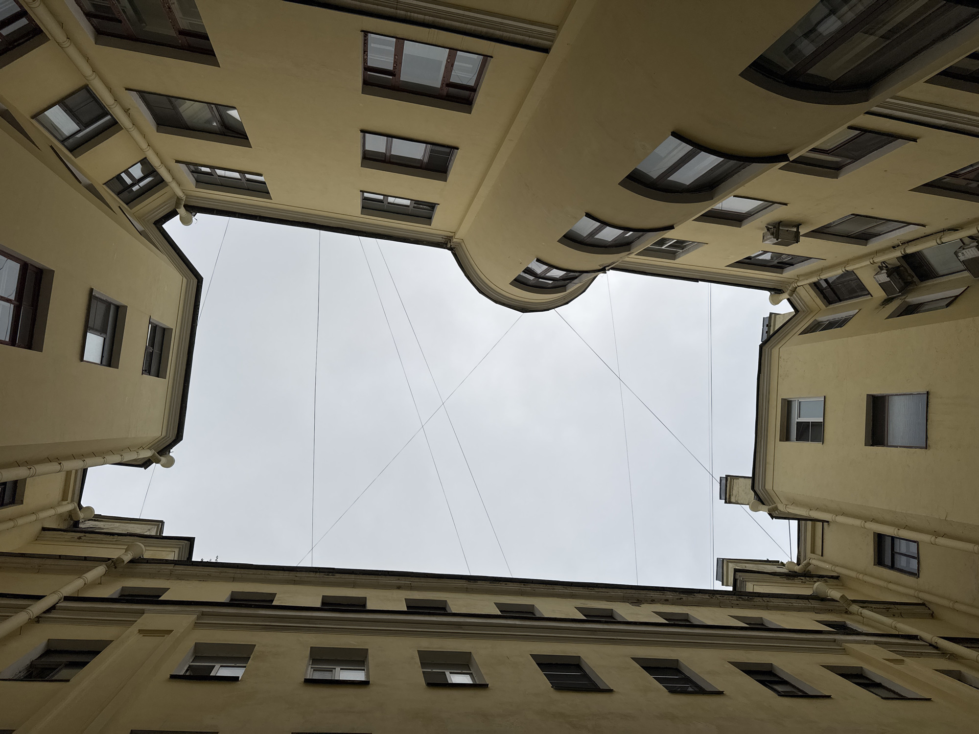 The image shows an upward view of the facades of old buildings in a courtyard in St. Petersburg, Russia. The buildings are made of yellow stucco and have multiple windows. The architecture suggests a pre-revolutionary era. Overhead, a grey, overcast sky is visible, crisscrossed by thin electrical wires. No people are present in the scene. The buildings appear to be residential or office blocks, typical of the historic city center. The perspective is dramatic, looking directly up from within the courtyard.