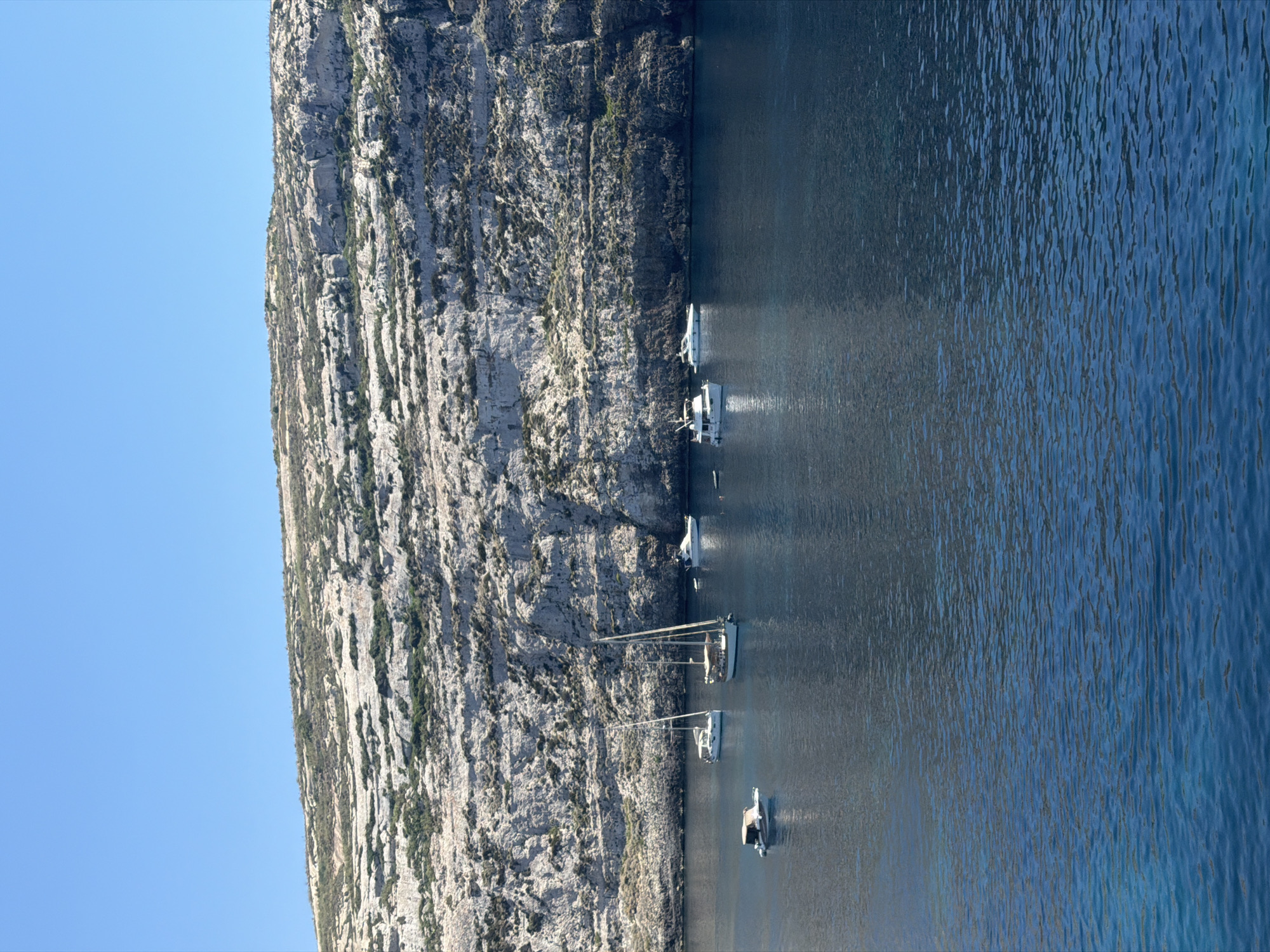 The image captures a scenic view of a rocky cliff face overlooking a calm, blue sea. Several boats are anchored in the water. In the foreground, a small white boat with a canopy is visible. Further back, a larger sailboat with two masts is moored, and next to it, another sailboat with a single mast. A white motorboat is positioned to the right of the sailboats. In the distance, along the base of the cliff, another white motorboat is seen. The cliff is characterized by its layered, light-colored rock and sparse green vegetation. The sky above is clear and bright blue. The location is identified as the coast of Saint Lawrence, Malta.