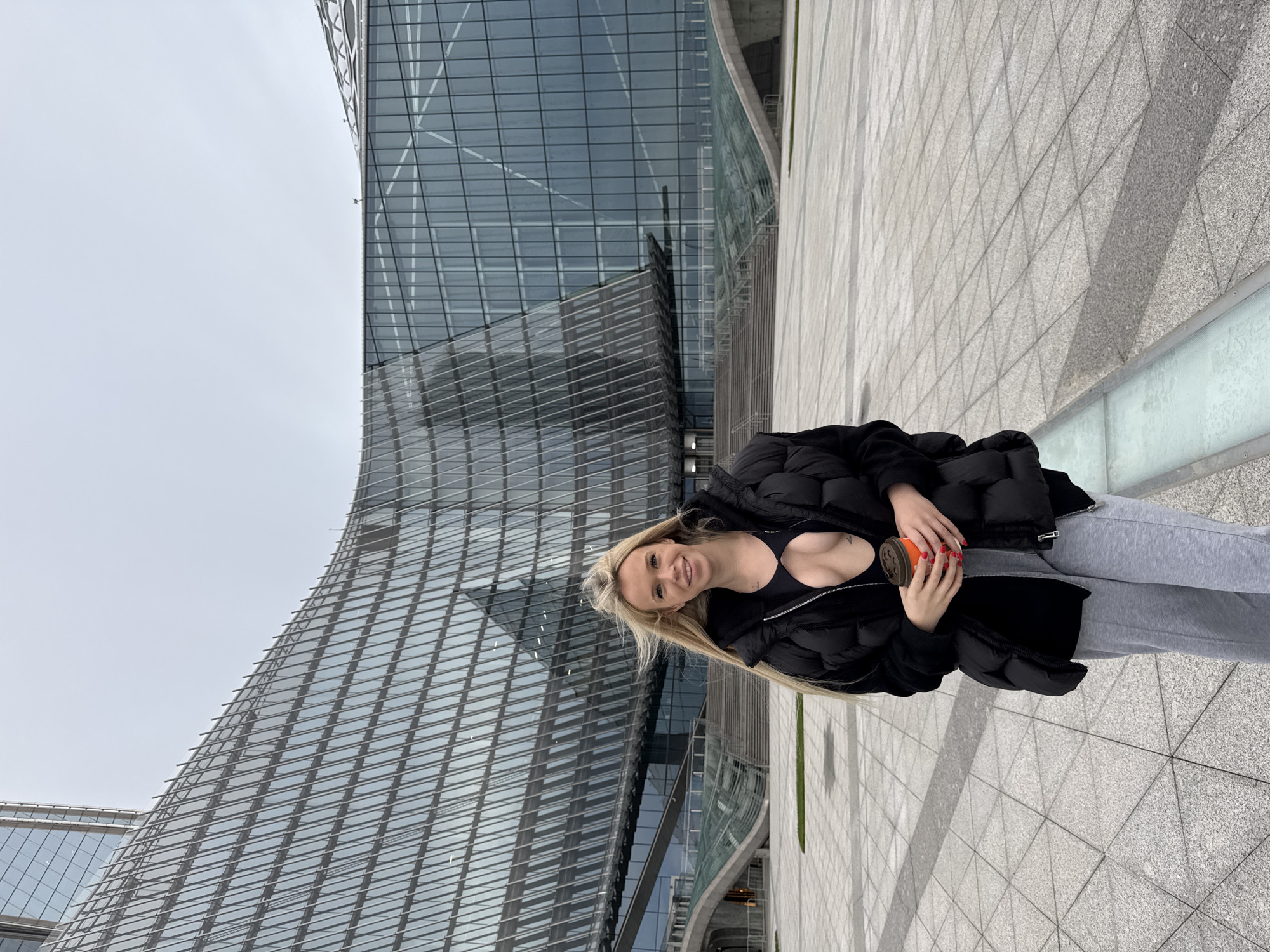 A young woman with blonde hair and a black puffer jacket stands in a plaza in front of a modern, curved glass building. She is wearing a black crop top and grey sweatpants, and is holding a takeaway coffee cup. Her expression is a smile, and her body language suggests she is relaxed. The building's facade is made of glass panels with a geometric grid pattern. The plaza is paved with large, light grey tiles arranged in a diamond pattern, with some green landscaping visible in the background. The sky is overcast, suggesting a cool or cloudy day. The architecture and paving hint at a contemporary urban environment, possibly a business district or a public space within a city. The time of day is not explicitly indicated but the lighting suggests daylight. The location cues, like the building style and paving, are consistent with modern architecture found in many large cities, including Saint Petersburg, Russia. There is no visible text in the image.