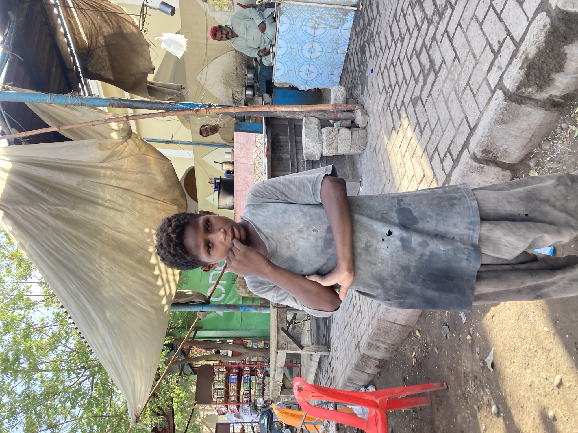 This vertical image captures a lively street scene in Uthal, Pakistan, under bright daylight with visible shadows. The central subject is a young boy with dark, curly hair and a serious expression, looking directly at the camera. He wears a plain, dark grey t-shirt and matching trousers, with one hand raised to his mouth.
Behind the boy, the scene unfolds into a bustling outdoor commercial area, characterized by informal structures and activity. Overhead, various canvas and tarpaulin sheets provide shade, supported by metal poles and wooden beams. To the left, lush green tree foliage is visible against the clear sky.
In the mid-ground, a makeshift shop or stall is evident, with shelves displaying numerous packaged goods, hinting at a small grocery or snack vendor. Partially visible behind these shelves is a green sign with unreadable white text. A red plastic chair is seen on the dirt ground in the lower right.
Further into the background, two adult men are present. One man, wearing a light-colored garment and a red cap, is seated behind a counter adorned with blue and white patterned tiles, suggesting a tea or food stall. Metal utensils and pots are visible on the counter. Another man's face is partially obscured but visible, observing the surroundings. The ground is a mix of light-colored paving stones and rough dirt, with some scattered debris. The overall impression is one of everyday life in a vibrant, informal marketplace.