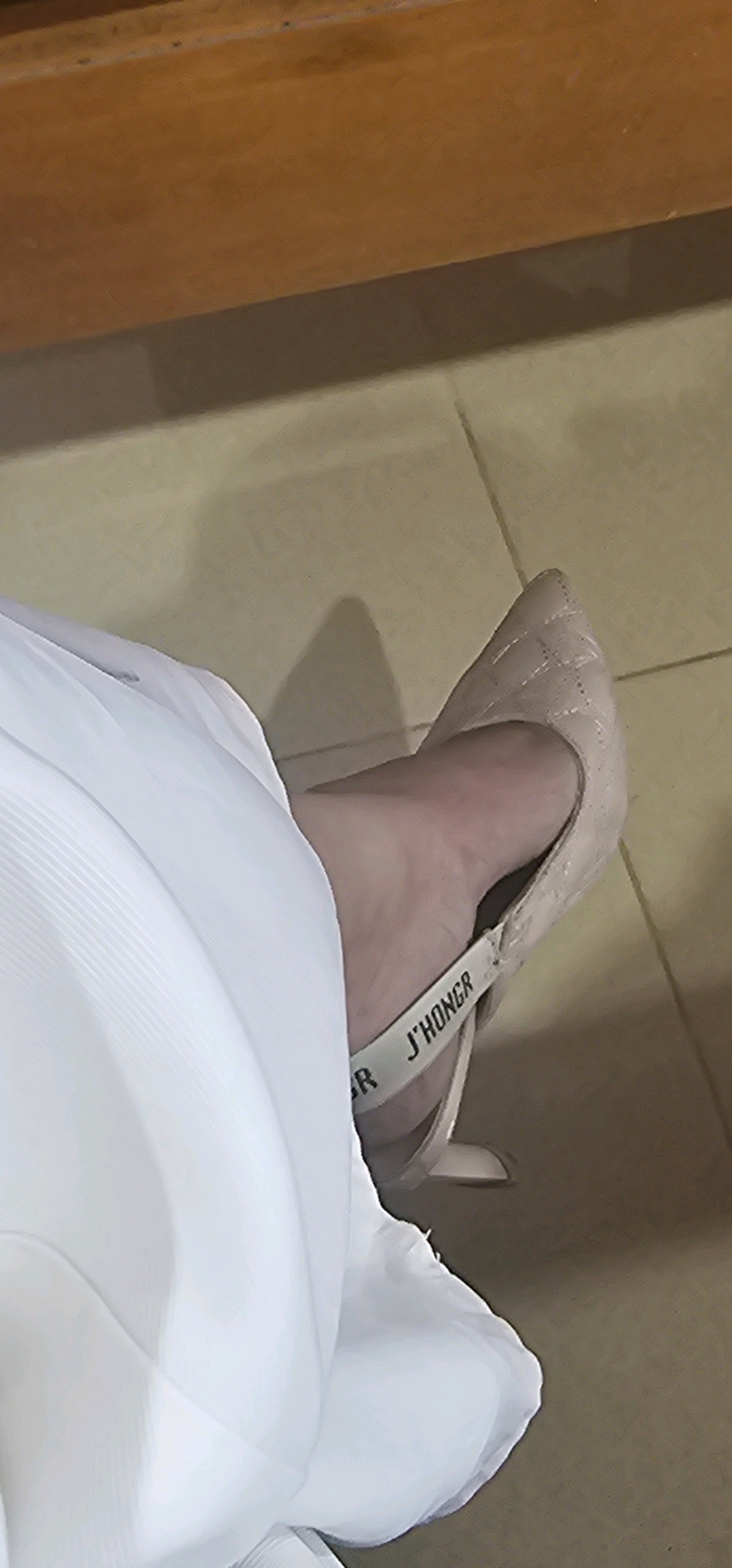 A close-up shot focuses on a person's foot wearing a light-colored, pointed-toe heel with a quilted texture. A white strap with black text, reading "R J'HONGR", is visible across the top of the foot. The person is wearing a white, flowing garment that drapes over the leg and shoe. The background consists of a tiled floor and a wooden surface above. The image does not provide any information about the location in Puncak Jaya, Indonesia.