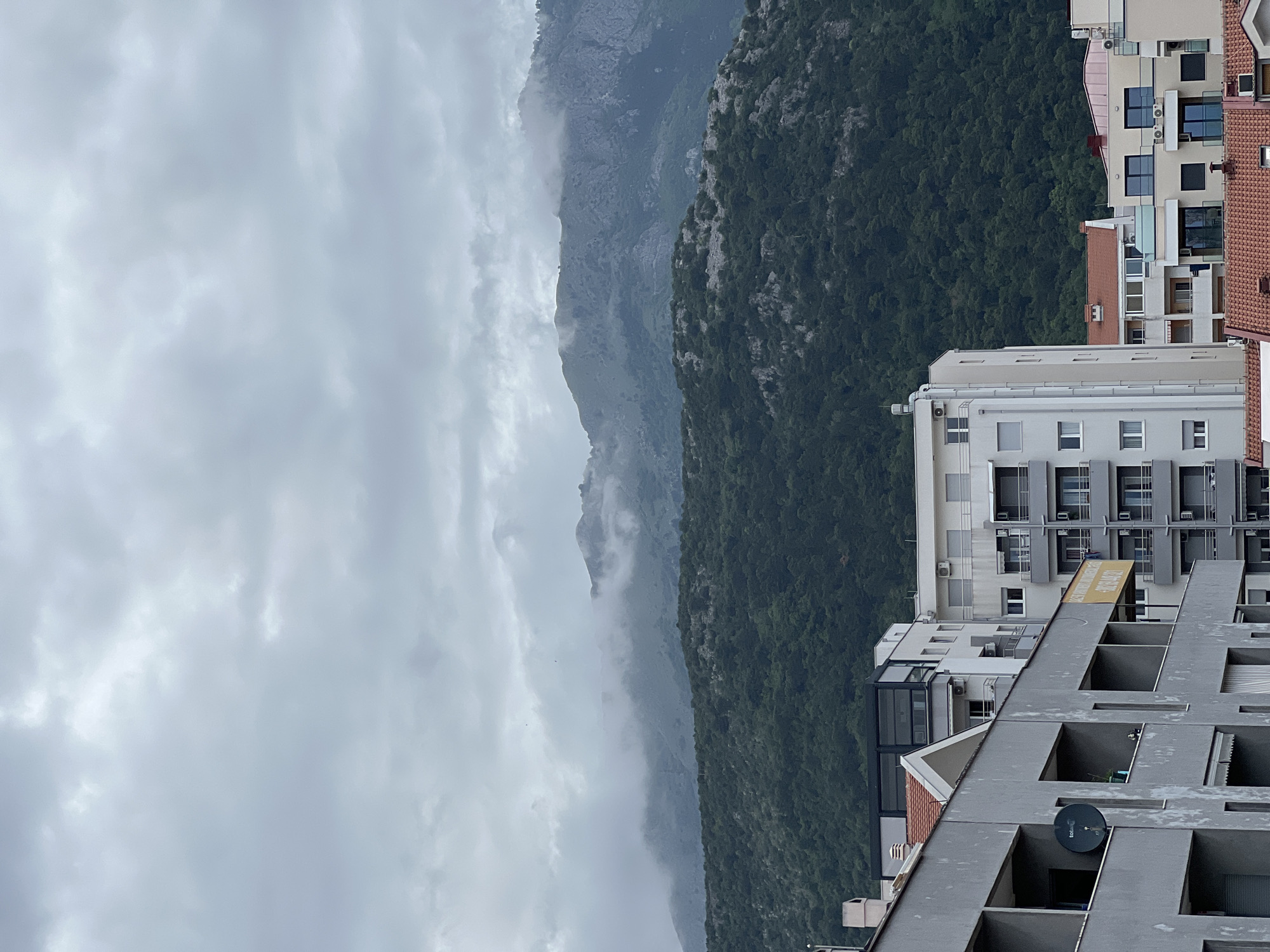 The image depicts a cityscape in Budva, Montenegro, viewed from an elevated position. A mountainous landscape, partially obscured by mist and clouds, forms the backdrop. In the foreground, multi-story residential buildings with a mix of architectural styles dominate the scene. The sky is overcast and gray. No people are visible. A satellite dish is attached to the building in the foreground.