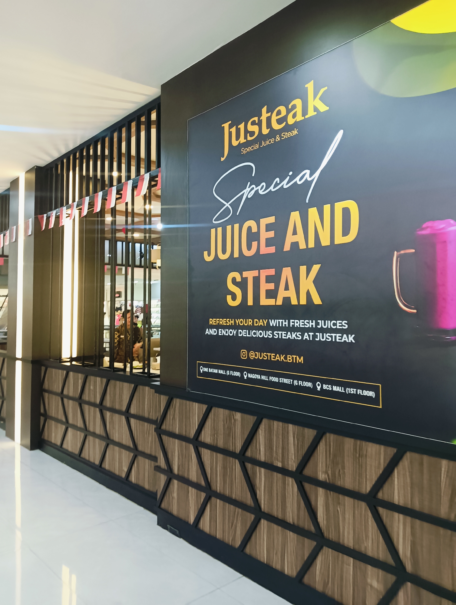 The image shows a close-up view of a storefront advertising "Justeak Special Juice & Steak". The advertisement is a large black banner with yellow and white lettering. The text prominently displays "JUICE AND STEAK" in large yellow letters, with "Special" written in cursive white font above it. Below this, it reads "REFRESH YOUR DAY WITH FRESH JUICES AND ENJOY DELICIOUS STEAKS AT JUSTEAK". A social media handle "@JUSTEAK.BTM" is visible.
The bottom of the banner lists several locations, indicating this business operates in Batam, Indonesia: "ONE BATAM MALL (G FLOOR)", "NAGOYA HILL FOOD STREET (G FLOOR)", and "BCS MALL (1ST FLOOR)".
The storefront features a dark wood-paneled facade with a geometric pattern of black lines. Small Indonesian flags are strung along a railing to the left of the banner. A person is partially visible through the railing, appearing to be an employee at a counter. The overall impression is a clean, modern commercial establishment.