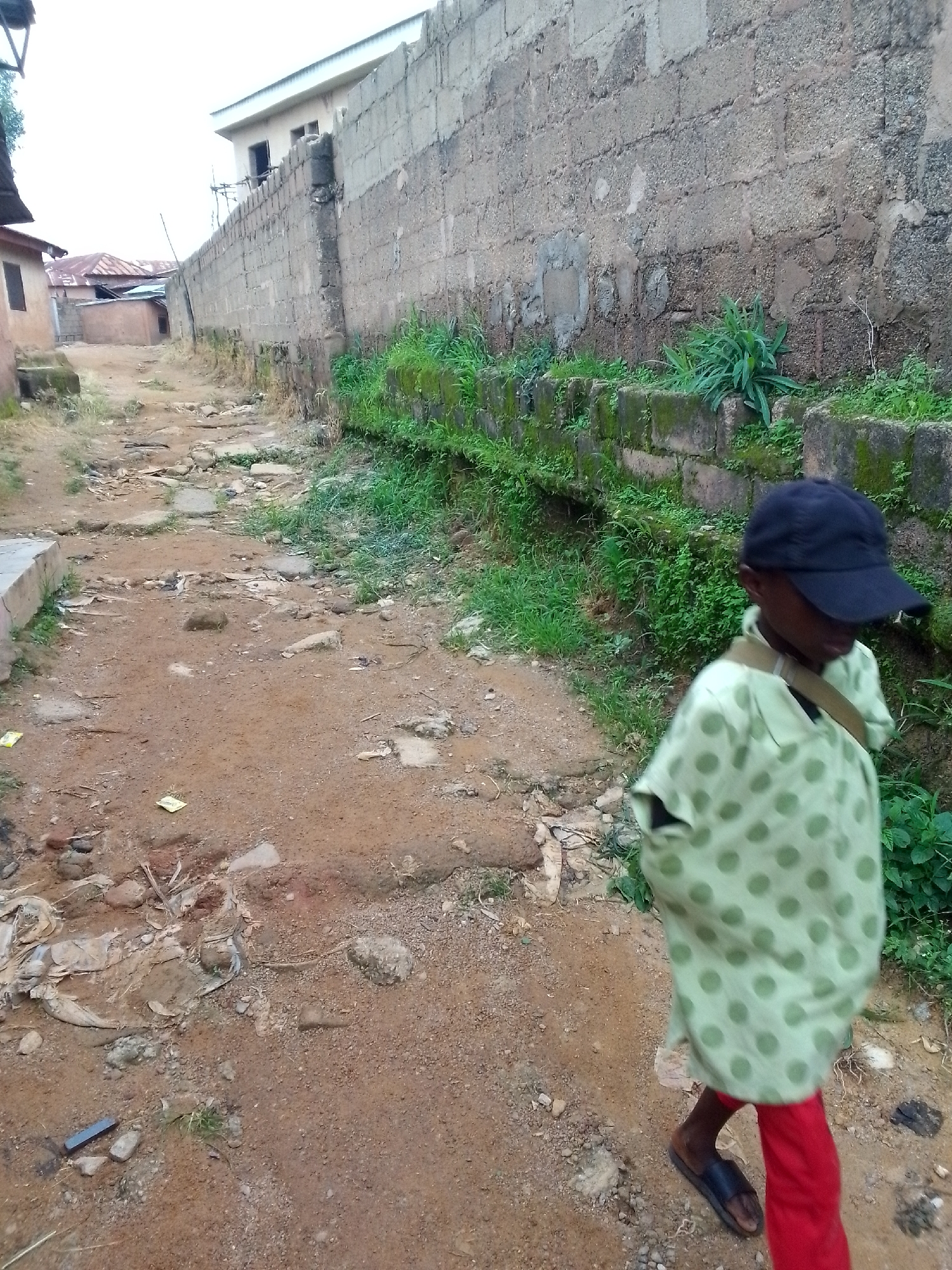 A young person wearing a light green, polka-dotted shirt, red pants, black sandals, and a black baseball cap walks along a dirt path in an urban area. The path is uneven and appears to be a dirt road with some scattered rocks and debris. A weathered concrete wall runs along the right side of the path, with overgrown green vegetation growing along its base and in patches on the wall itself. In the background, buildings with red roofs are visible, suggesting a residential or commercial area. The sky is overcast. The scene appears to be in a developing or rural part of a city.