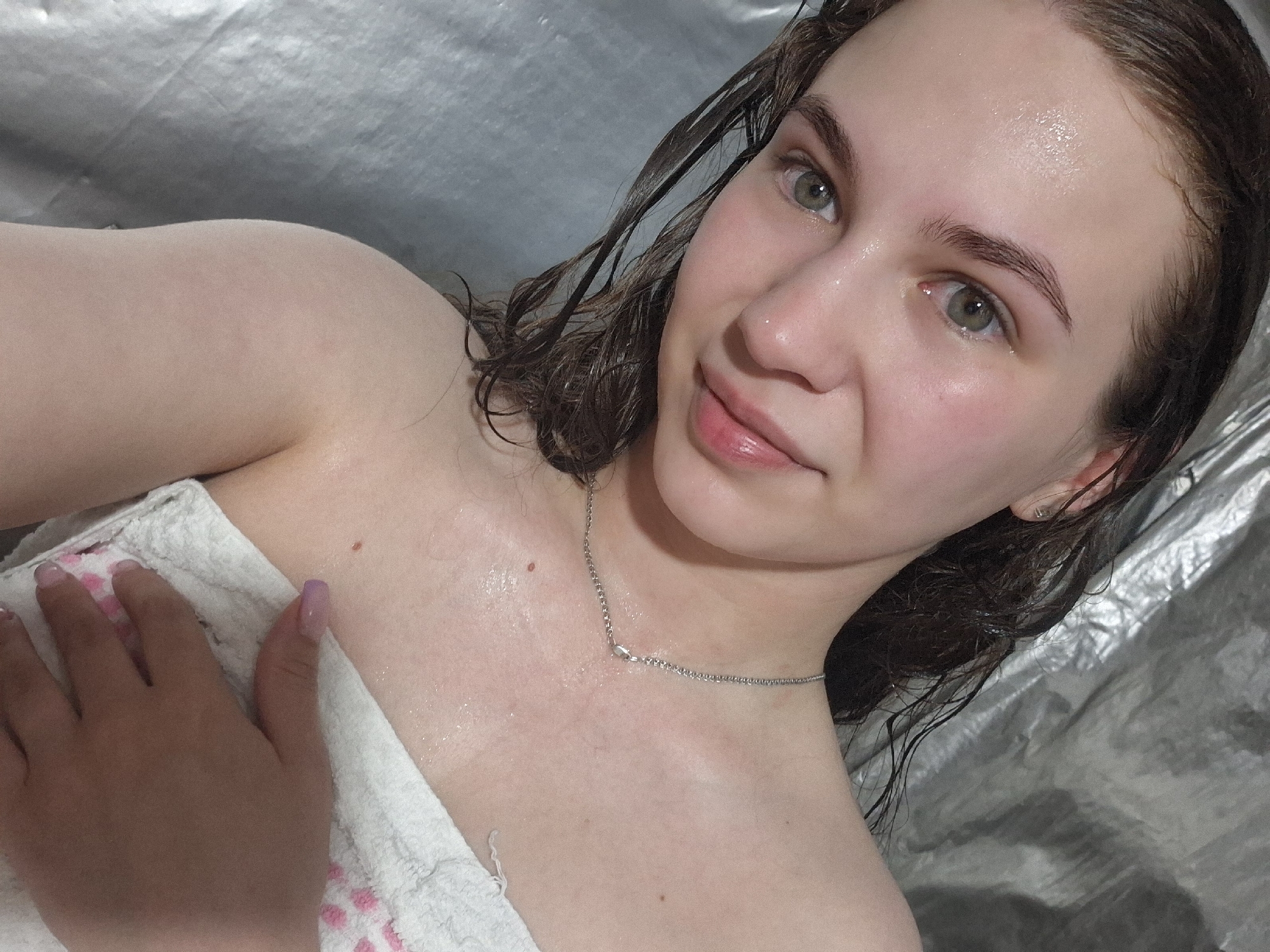 A medium shot captures a woman with wet hair, possibly after a shower, as evidenced by the glistening skin and damp hair. She is partially covered by a white towel with pink accents. A delicate silver necklace adorns her neck. Her nails are manicured with a light purple color. The background is a crumpled, reflective silver material. The location is not discernible from the image.