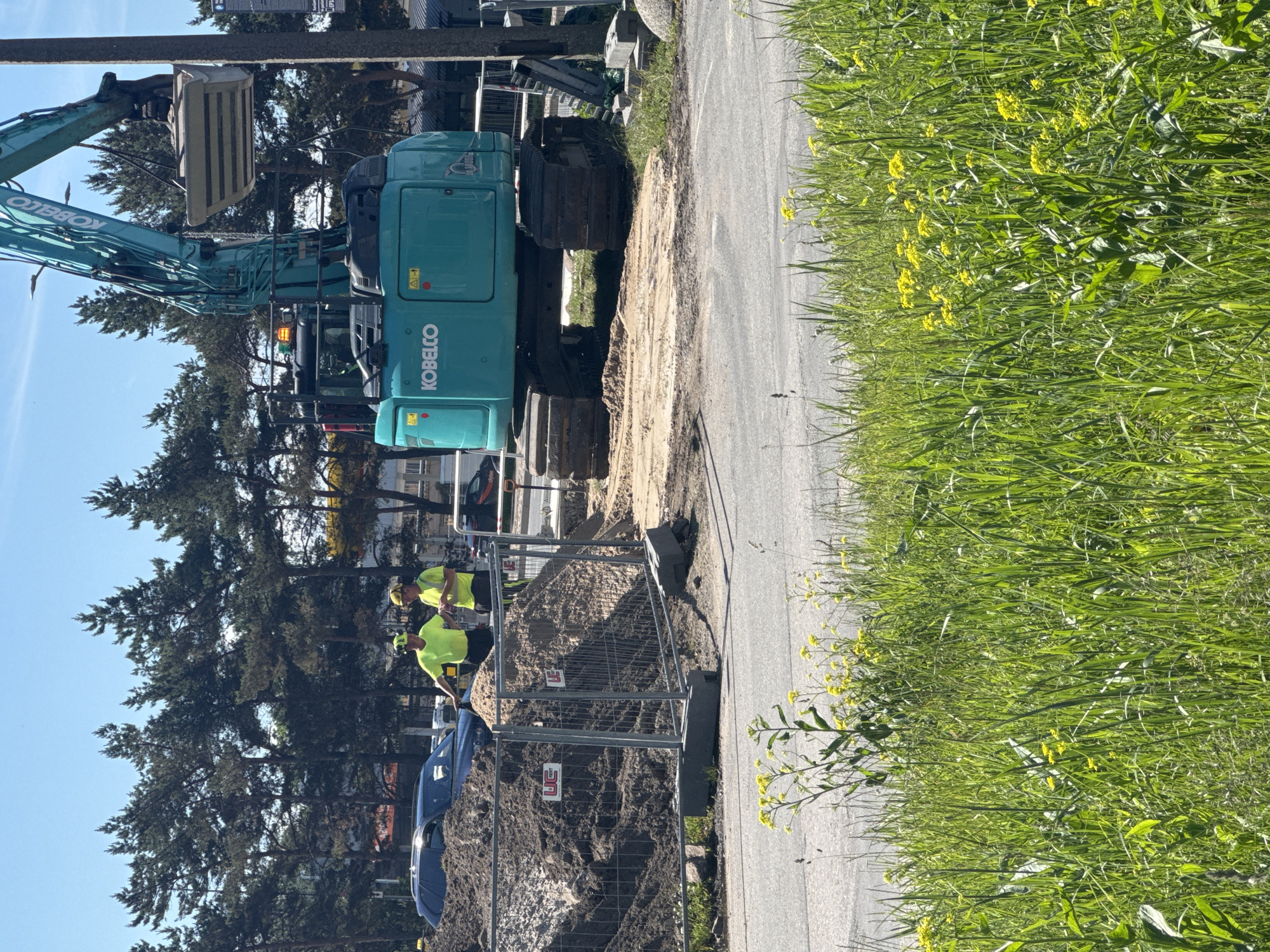 A bright, sunny day in Tallinn, Estonia, shows a construction site with an excavator. Two construction workers in yellow high-visibility vests and hard hats stand near a pile of dirt. One worker is leaning on the dirt pile and looking at the other worker, who is holding a phone. A large, turquoise Kobelco excavator is positioned on the site. In the background, pine trees line the perimeter of the construction area, and a blue car is parked near the dirt pile. The foreground of the image is filled with lush green grass and yellow wildflowers.