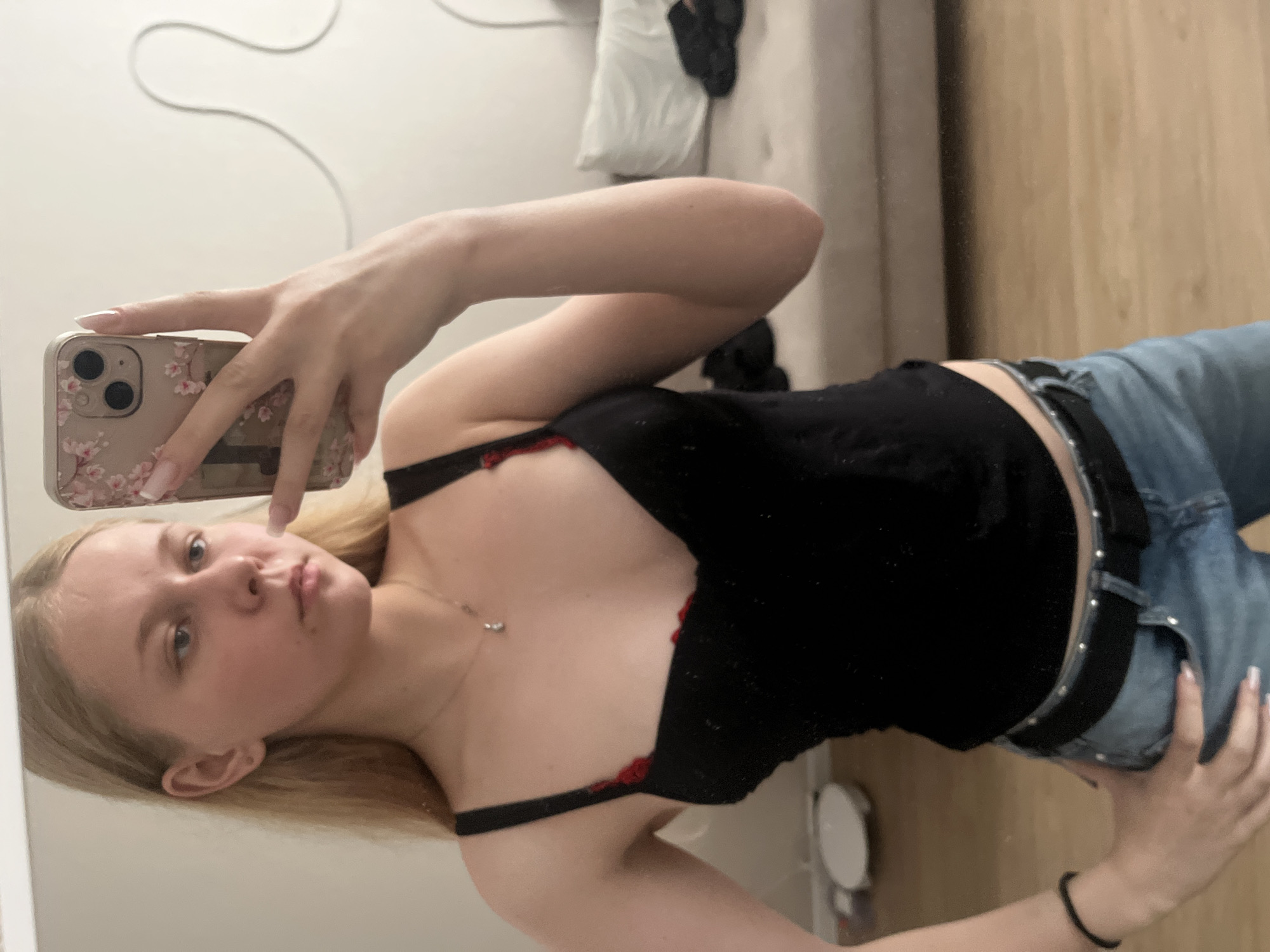 The media file is a portrait of a young Caucasian woman with blonde hair, taken from the chest up. She is holding a smartphone in her right hand, with a floral-patterned case, and appears to be taking a selfie in a mirror. The woman is wearing a black top with red accents and blue jeans. She has light skin and her expression is a playful pout. A delicate silver necklace with a small pendant is visible around her neck.
The background suggests an indoor setting, possibly a bedroom or dressing area, with a light-colored wall and a wooden surface that could be furniture or a wall panel. A white object, possibly clothing, is hanging in the background. The lighting suggests it is daytime. There is no visible text in the image. The image is oriented vertically.