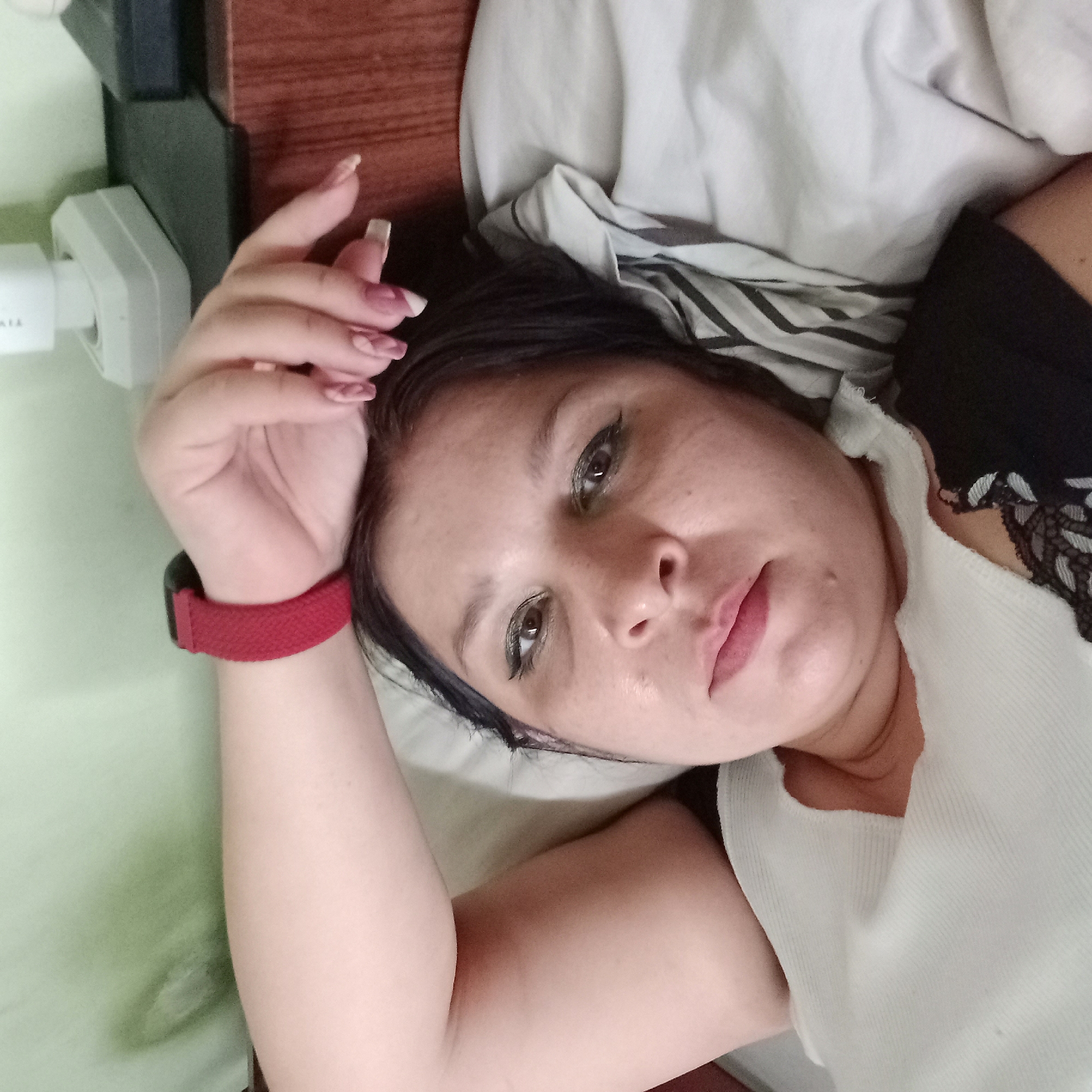 A woman is lying in bed, her head resting on her arm. She is wearing a white top and has a red watch on her wrist. The room appears to be a bedroom with a wooden headboard and a light green wall. The woman's eyes are open, and she is looking at the camera. There is a power outlet visible on the wall behind her. The location is not specified in the image or the provided text.