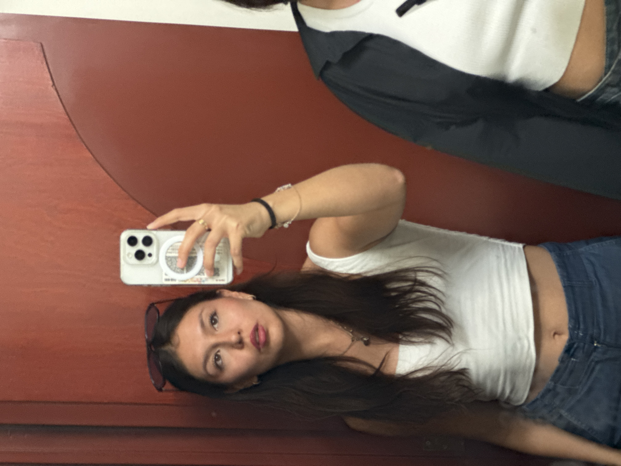 A young woman is depicted taking a mirror selfie, with the image rotated 90 degrees counter-clockwise. She is the central subject, featuring long dark hair and wearing a white sleeveless crop top that exposes her midriff, along with blue denim jeans. Sunglasses are perched on her head, and she wears a bracelet on her left wrist and a ring on her right hand. She holds a light-colored smartphone, likely an iPhone due to its visible multi-camera array, which has a white circular grip or pop socket accessory on its back. Her lips are slightly puckered, and she gazes directly into the camera with a casual expression.
Partially visible above her in the reflection is another person's torso, clad in a white top beneath a dark jacket or shirt. The selfie is taken indoors against a reddish-brown wall or door, with a strip of white visible at the top of the background, likely indicating a ceiling or trim. While a pattern resembling a QR code or text is visible on the phone's accessory, it is not legible. The overall activity is a self-portrait taken in a reflection.