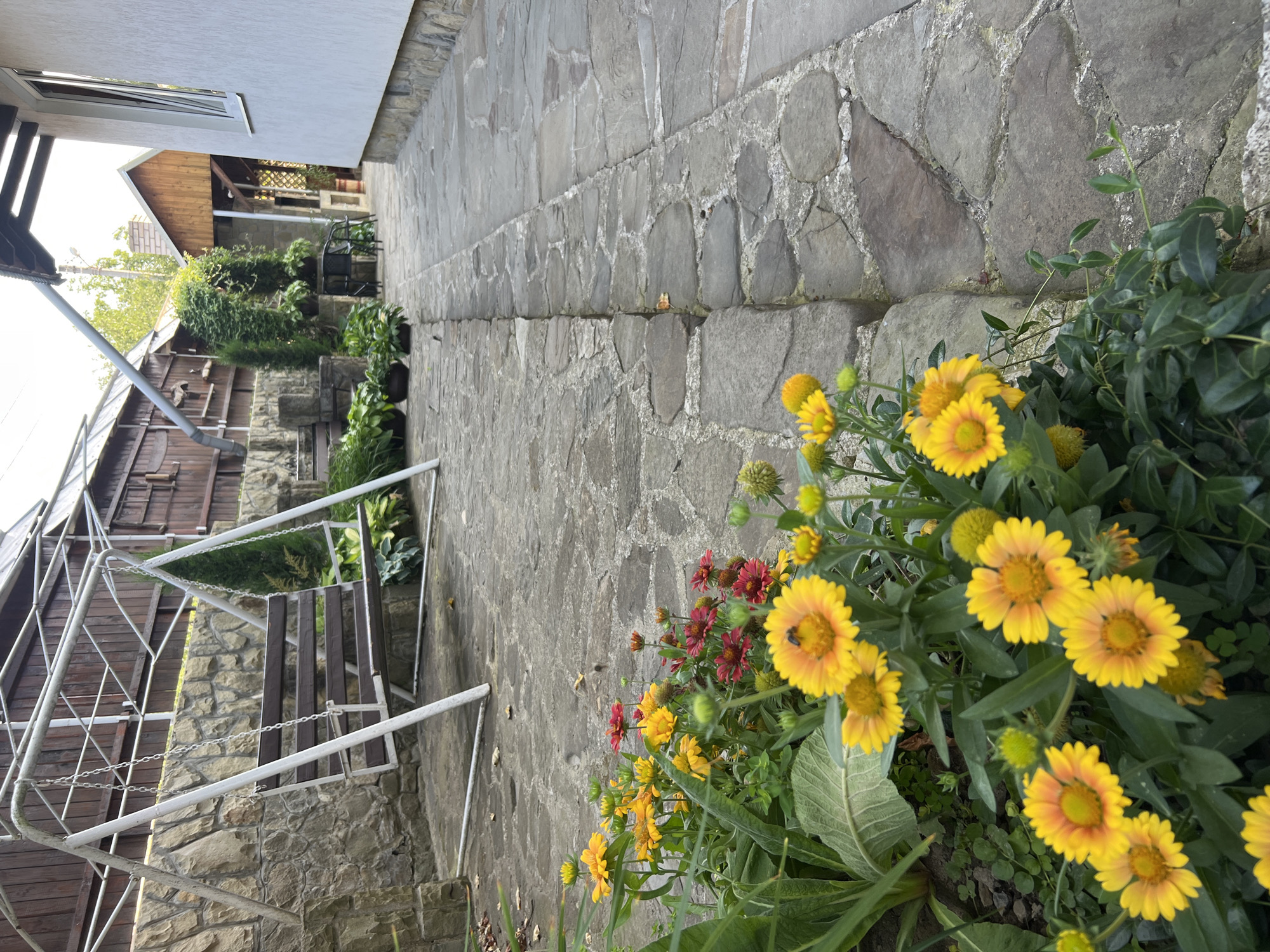 In Yaremche, Ukraine, a stone pathway leads past a garden bed filled with blooming yellow and red flowers. A white metal swing set with a dark wooden seat stands on a stone patio to the left, adjacent to a stone wall. Behind the patio, a wooden fence encloses the area. Further back, a building with a wooden roof and a white facade is visible, with an open doorway revealing a dark interior. The scene suggests a private backyard or garden setting.