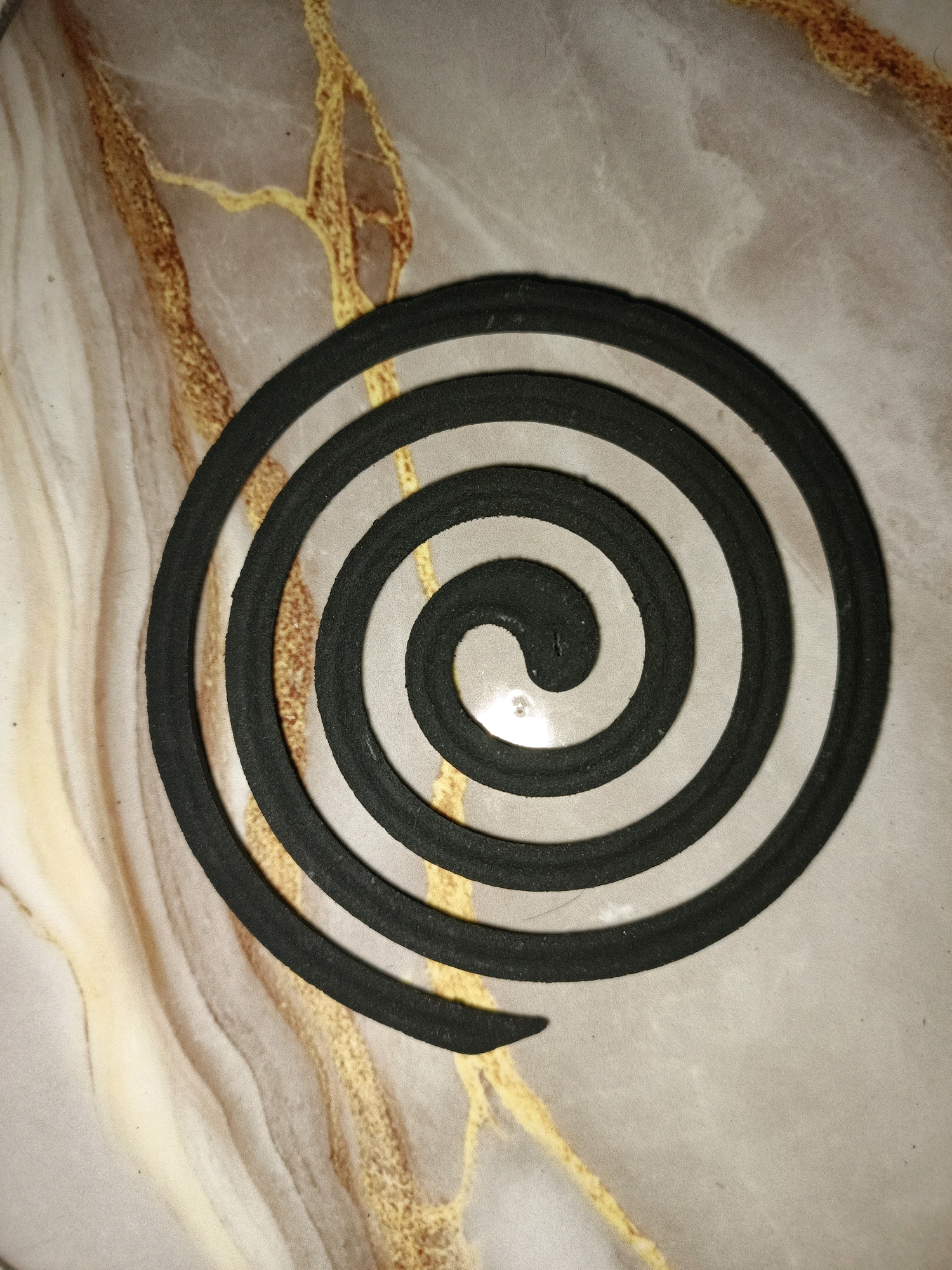 A black mosquito coil is centrally placed on a light-colored, marbled surface with gold-colored veins. The coil is in a spiral shape and appears to be made of a dark, granular material. The surface beneath it exhibits a pattern reminiscent of polished stone or resin, with swirling light beige and golden brown lines. The lighting creates subtle shadows around the coil, highlighting its texture. The scene is photographed from a top-down perspective. There are no people or actions depicted in the image. The specific location in Ketegan, Indonesia, is not identifiable from the visual information provided.