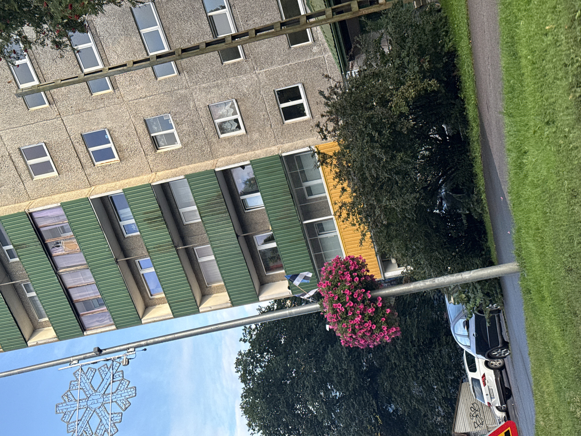 The image shows a residential building in Tallinn, Estonia, with a grey concrete facade and green balconies. A large, illuminated snowflake decoration hangs from a pole on the left side of the image. A pot of pink flowers is attached to the same pole. Two flags, one a national flag and the other with horizontal stripes of blue, black, and white, are also attached to the pole. In the foreground, a paved road runs alongside a grassy area. Several cars are parked on the side of the road, and lush green trees and bushes are visible throughout the scene. The sky is blue with some clouds.