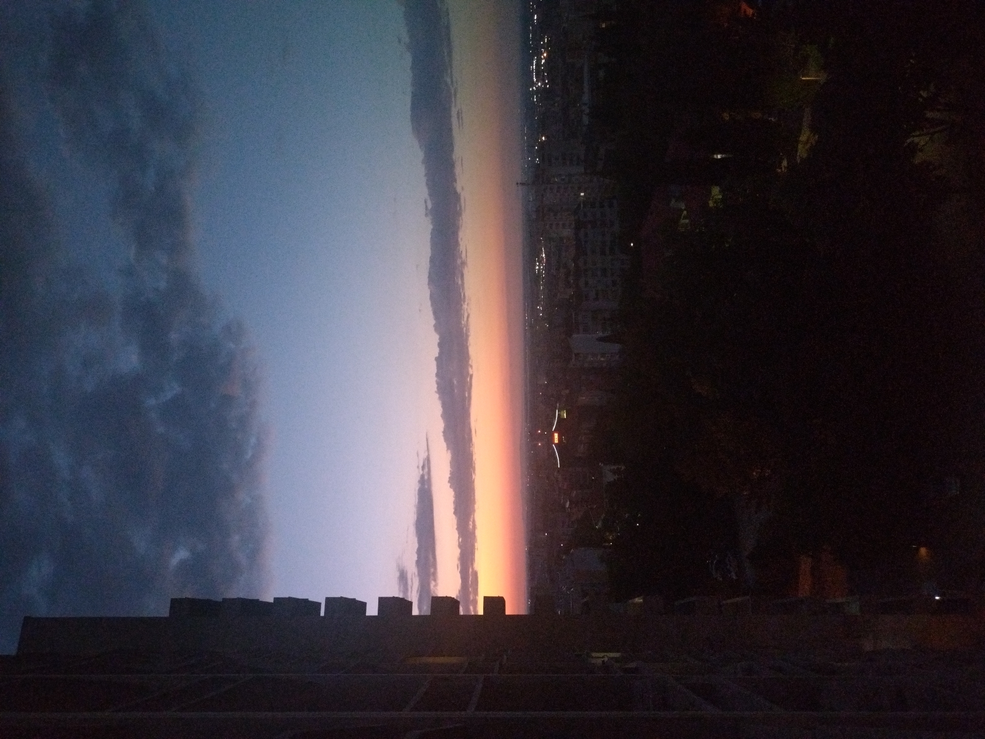 The image captures a twilight view of Stara Zagora, Bulgaria, from an elevated position. In the foreground, the dark, unfinished side of a building, possibly under construction, dominates the left frame, its balconies creating a repeating geometric pattern. The sky transitions from deep blue and grey clouds at the top to a soft gradient of pink, orange, and pale blue near the horizon. Below the sky, the cityscape of Stara Zagora is visible, characterized by rows of buildings, some with illuminated windows, and the faint glow of streetlights. Dark foliage is interspersed throughout the middle ground, obscuring parts of the lower city. The overall impression is that of a city at dusk, viewed from a high vantage point overlooking residential and urban areas.