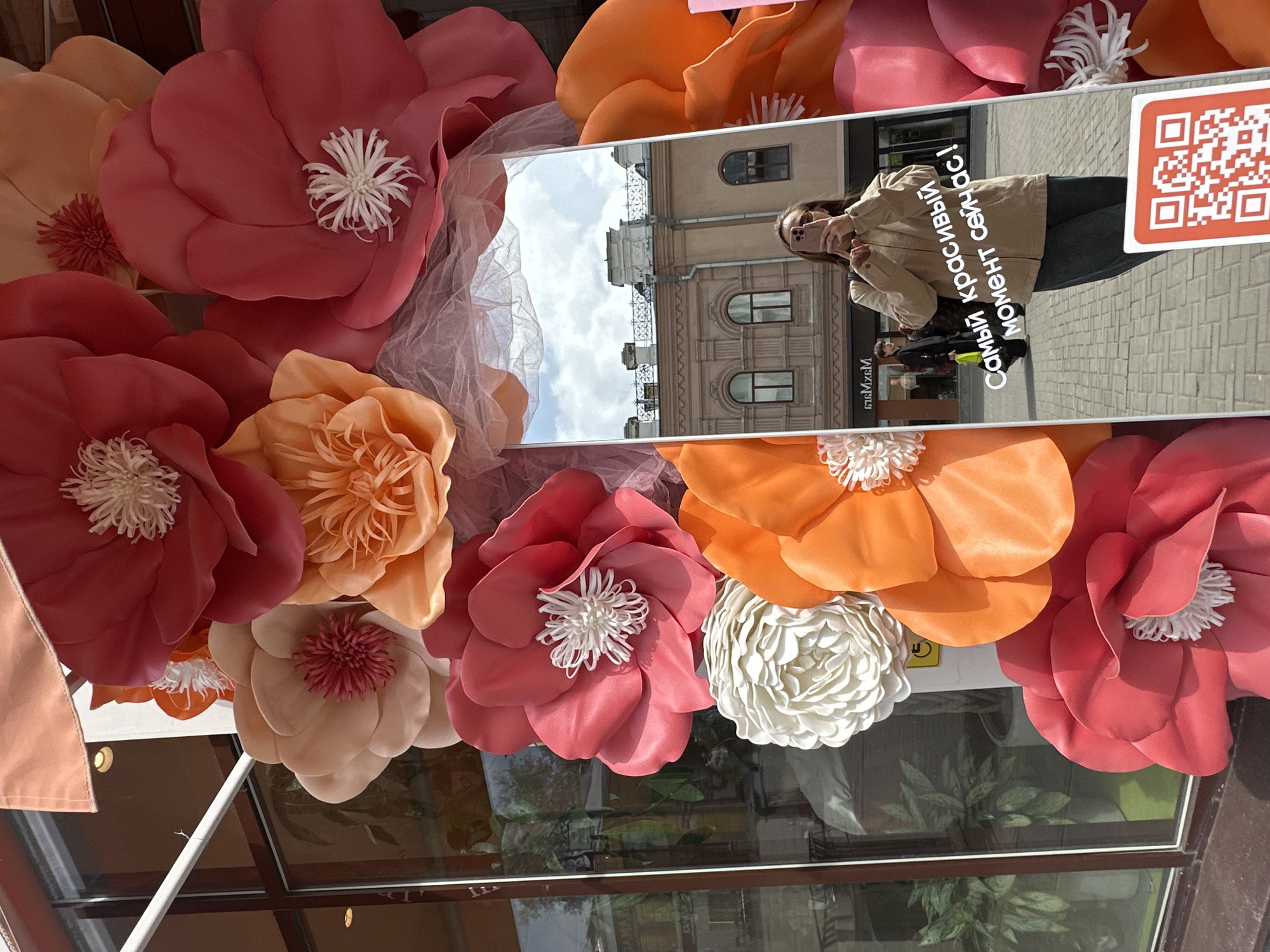 The scene shows an eye-level, medium shot of a shop window display decorated with oversized paper flowers in shades of peach, orange, red, and white. A rectangular mirror is centrally positioned among the flowers, reflecting a young woman taking a selfie with her phone. The reflection also includes a building exterior with arched windows and a light blue sky with clouds. In the lower part of the mirror's reflection, other pedestrians are visible. Cyrillic text and a QR code are present on the right side of the mirror's reflection. Through the lower part of the window, plants and reflections of the outside environment are visible. Based on the Cyrillic text and the QR code information, the scene is likely located in Yekaterinburg, Russia.