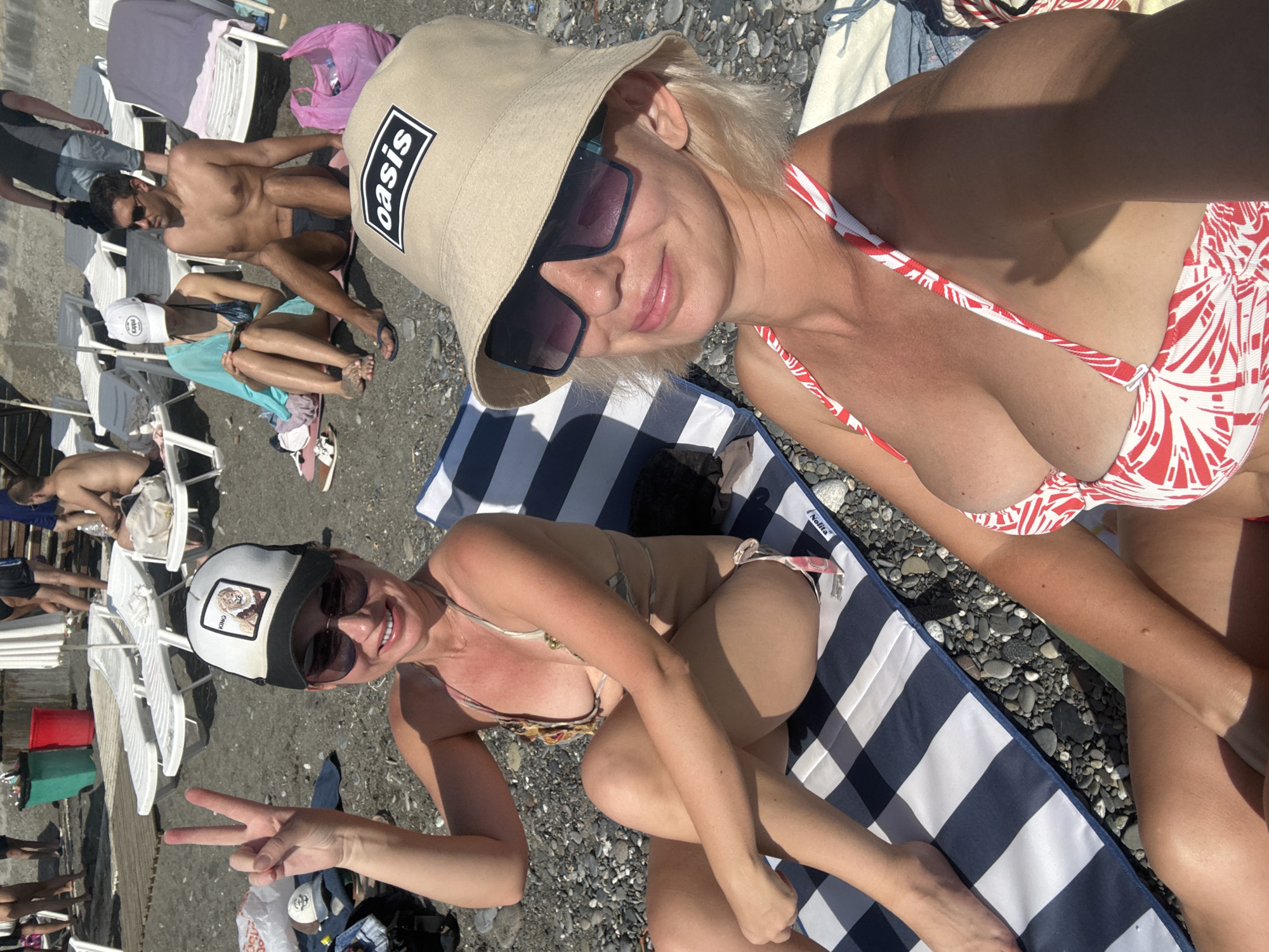 This is a medium shot, eye-level, full color, outdoor photograph taken on a sunny day at a beach in Sochi, Russia. In the foreground, two women are sitting on a striped beach towel on a pebble beach. The woman on the left is wearing sunglasses and a baseball cap with a lion logo. She is holding up two fingers in a peace sign and smiling at the camera. She is wearing a patterned bikini. The woman on the right is wearing a bucket hat with "oasis" printed on it and large sunglasses. She is also wearing a red and white patterned bikini. Her left arm is extended toward the camera. In the background, there are beach chairs, other people, and the sea. The overall tone of the image is casual and summery.
