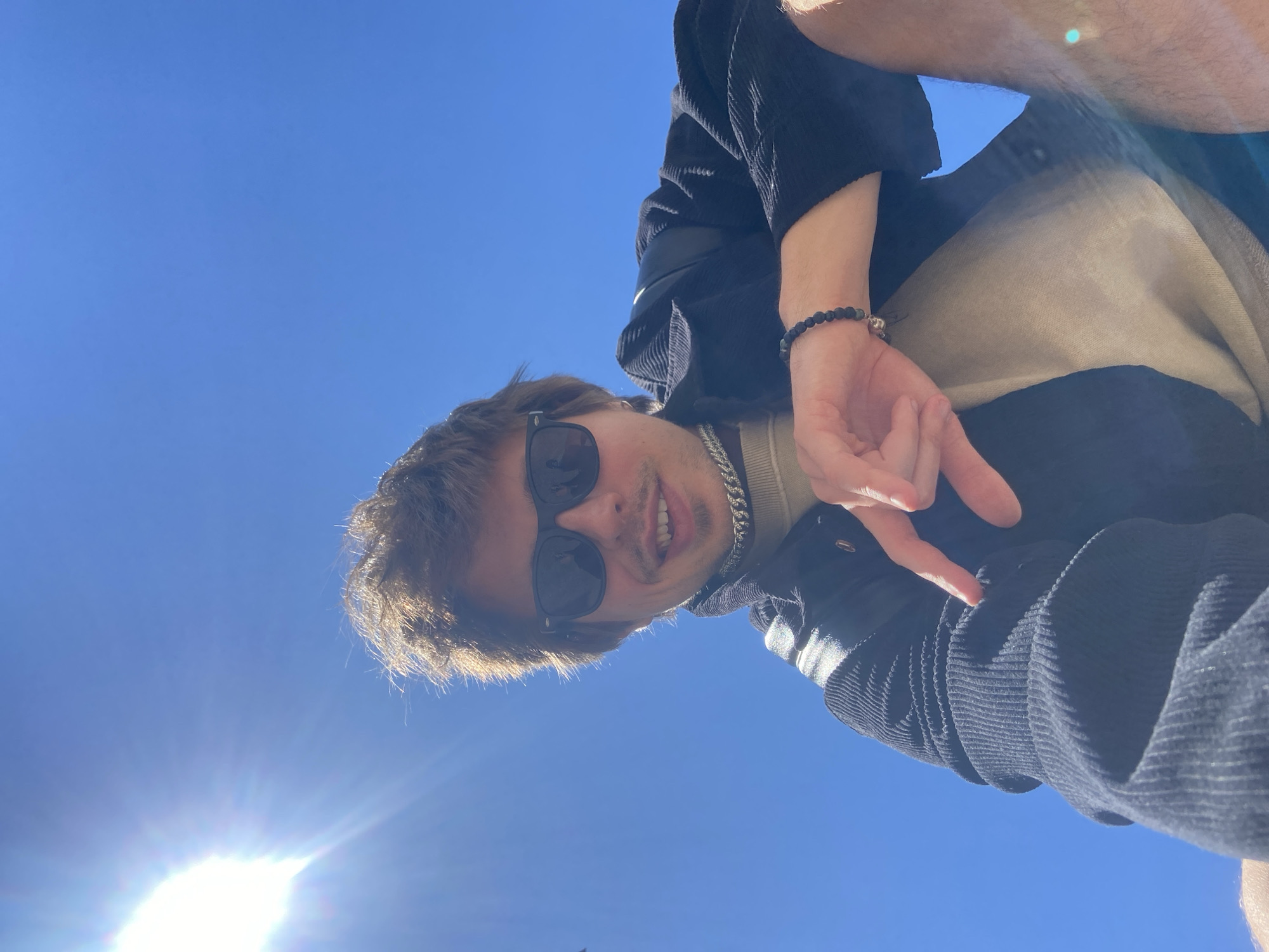 A person is seen from a low angle looking down toward the camera under a bright blue sky. The sun is shining brightly in the lower left corner of the frame. The person is wearing sunglasses and a chain necklace, with a dark corduroy jacket over a light-colored top. The person’s hand is visible near the top of the image, gesturing downward. The location, Šušanj, Montenegro, cannot be verified from the image content.