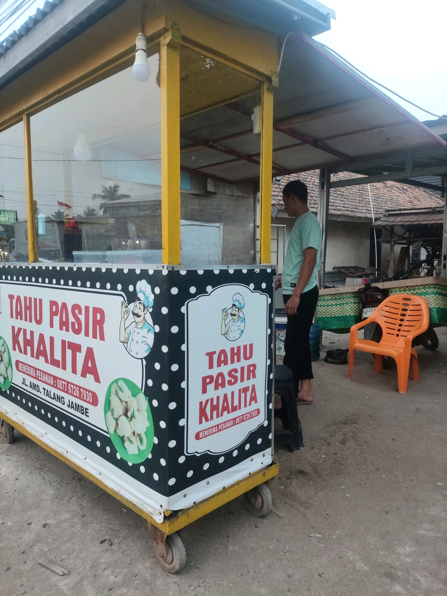 The image shows a mobile food cart selling "Tahu Pasir Khalita" in Palembang, Indonesia. The cart is yellow and white with a black and white polka dot pattern and graphics of a chef and the product. The cart has wheels and appears to be parked on a dirt or gravel surface. A man in a light green t-shirt and black pants is standing next to the cart. Behind the cart, there are buildings with tiled roofs and a motorcycle. An orange plastic chair is visible in the foreground. The overall scene suggests a street-side food vendor operating in a moderately urban or suburban environment.