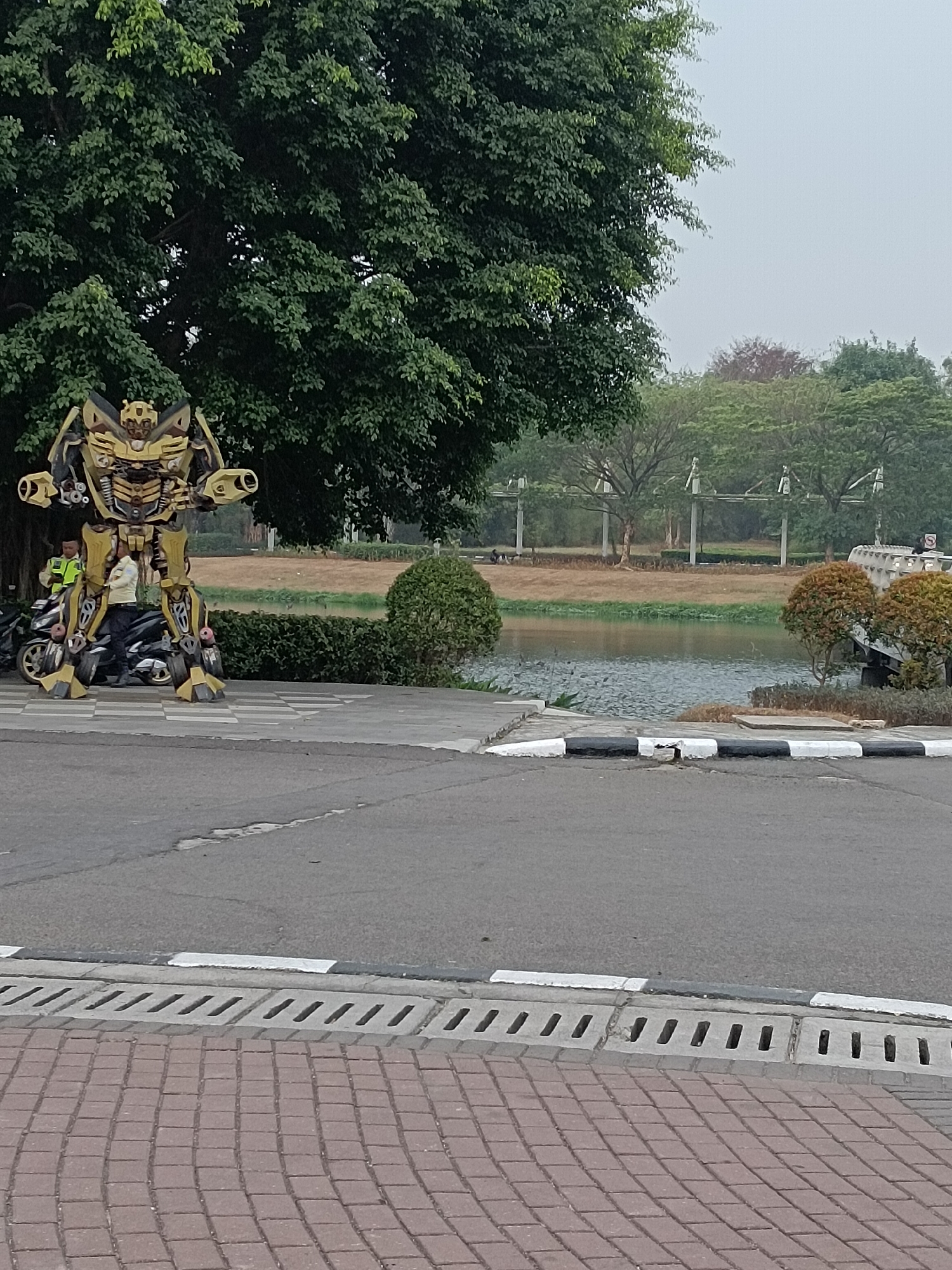 An outdoor scene in Jayamukti, Indonesia features a large, yellow and black robot statue resembling Bumblebee from Transformers. The robot stands on a paved walkway. To the left of the robot, two individuals are present, one wearing a yellow vest and standing next to a black motorcycle. The robot and motorcycle are situated near a large tree with dense green foliage. In the background, a body of water is visible, bordered by brown grassy areas and more trees. Further in the distance, a bridge-like structure with white pillars and a light-colored roof spans across the water. The ground in the foreground is paved with reddish-brown bricks, and a concrete drainage grate with a black and white striped curb runs along the edge of the asphalt road. The sky is overcast and light gray.