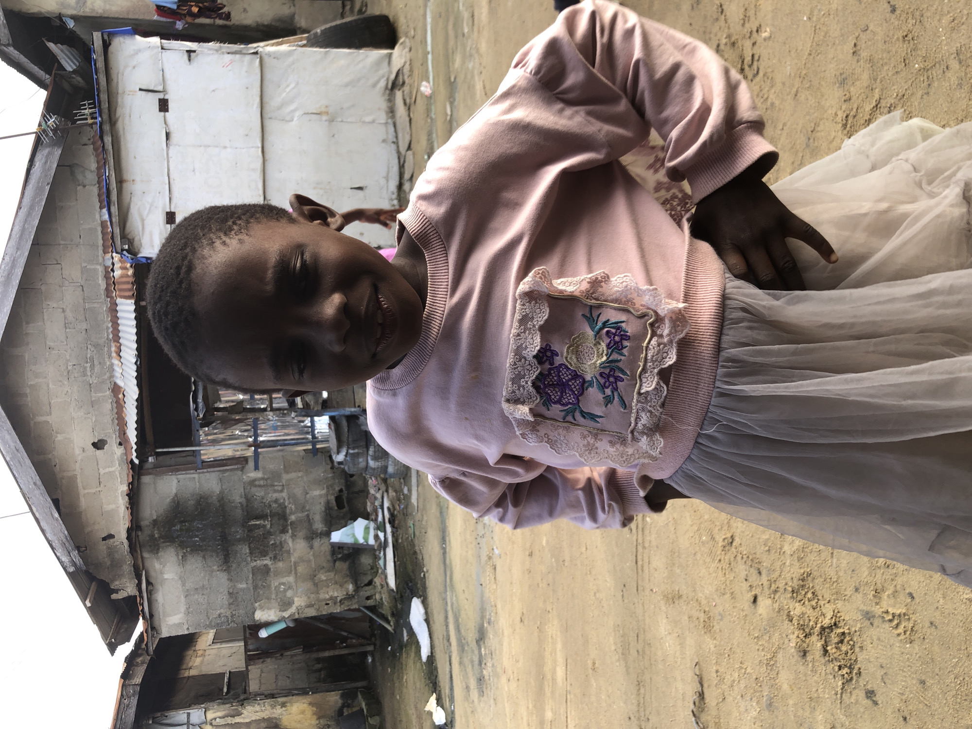 In Ado, Nigeria, a young girl stands in front of a dilapidated building. She is wearing a pink sweatshirt with a floral applique on the chest and a light grey tulle skirt. The building behind her appears to be constructed from cinder blocks and corrugated metal, with some sections in disrepair. The ground is a sandy dirt path. The lighting suggests it is daytime, possibly late afternoon due to the soft shadows. The overall scene depicts a humble, possibly impoverished, environment.