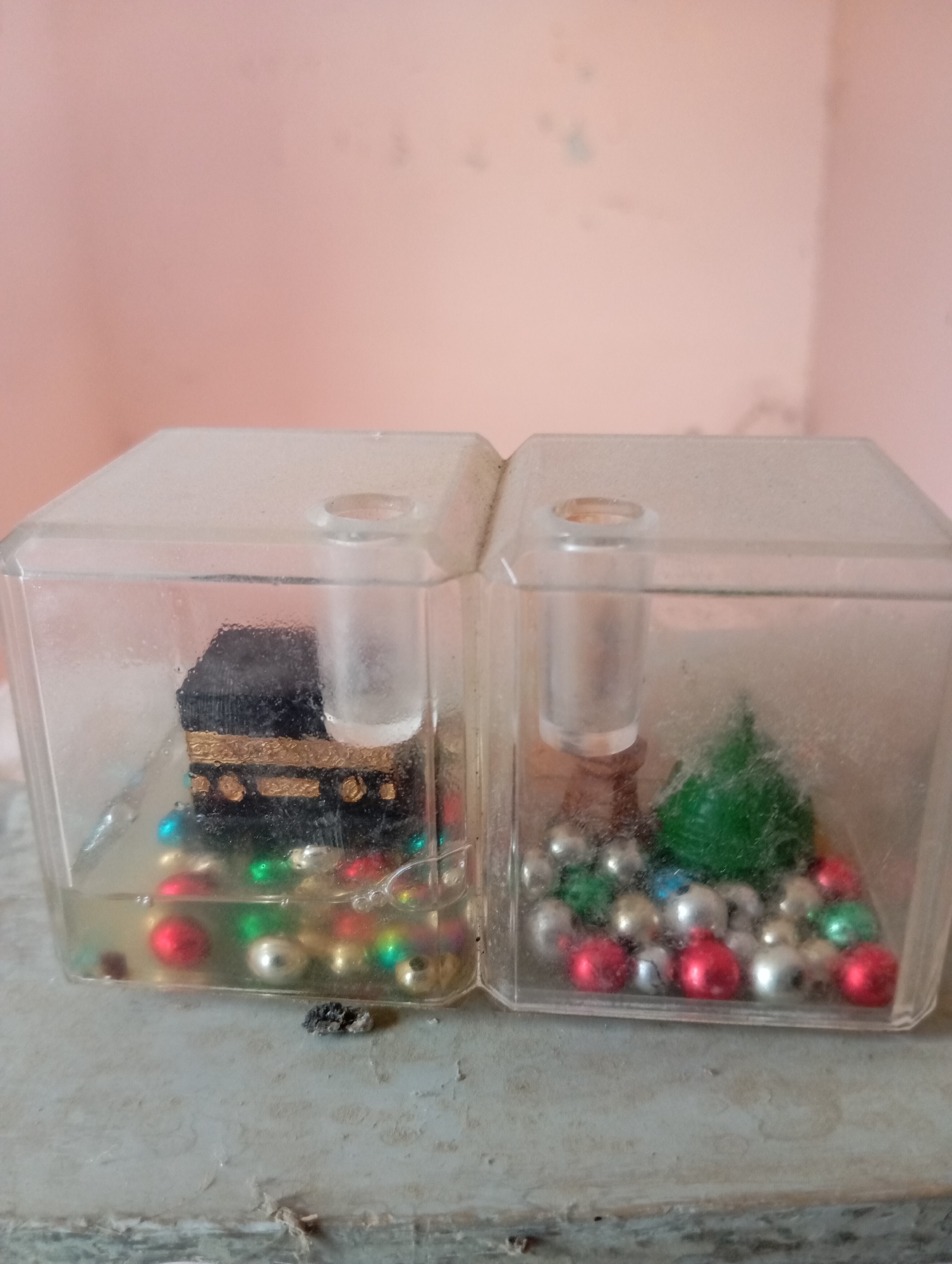 The image shows two small, clear plastic cube-shaped containers, each filled with a miniature scene. The left container holds a replica of the Kaaba, the Islamic holy site in Mecca, surrounded by colorful beads in liquid. The right container features a small green Christmas tree-like object surrounded by similar colorful beads, suggesting it might be a decorative piece. Both containers are placed on a dusty, textured surface against a light pink wall. The overall scene appears to be indoors, possibly on a shelf or table.