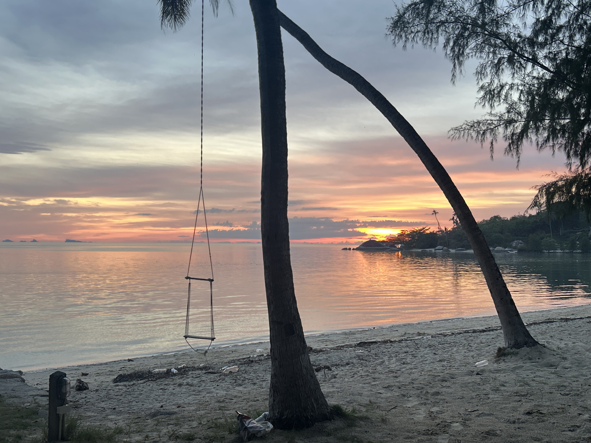 A serene beach scene at sunset in Ko Pha Ngan, Thailand. The sky is painted with soft hues of orange, pink, and purple, reflecting on the calm ocean water. Two palm trees stand on the sandy shore, one leaning dramatically to the right, with a swing hanging from its trunk. The beach itself is mostly bare sand, with some scattered debris, and a small light fixture on a wooden post on the left. In the distance, lush green islands dot the horizon. The overall atmosphere is peaceful and tranquil.