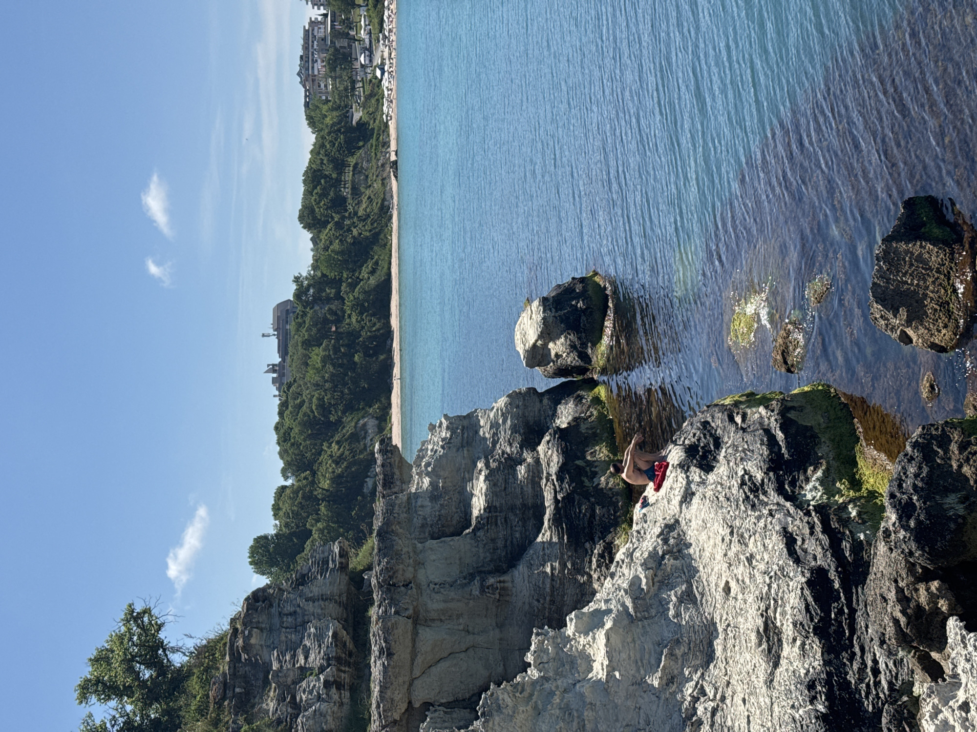 A person is sitting on a rocky outcrop overlooking a body of blue water. The person is wearing blue shorts and is shirtless, with a red towel or piece of clothing beside them. The rocky outcrop is part of a cliff face that extends to the left of the frame. The water is clear and shows submerged rocks and seaweed. In the background, a sandy beach with numerous umbrellas is visible, bordered by green trees and buildings. The sky is blue with a few scattered white clouds. The location is the coast of Tsarevo, Bulgaria.
