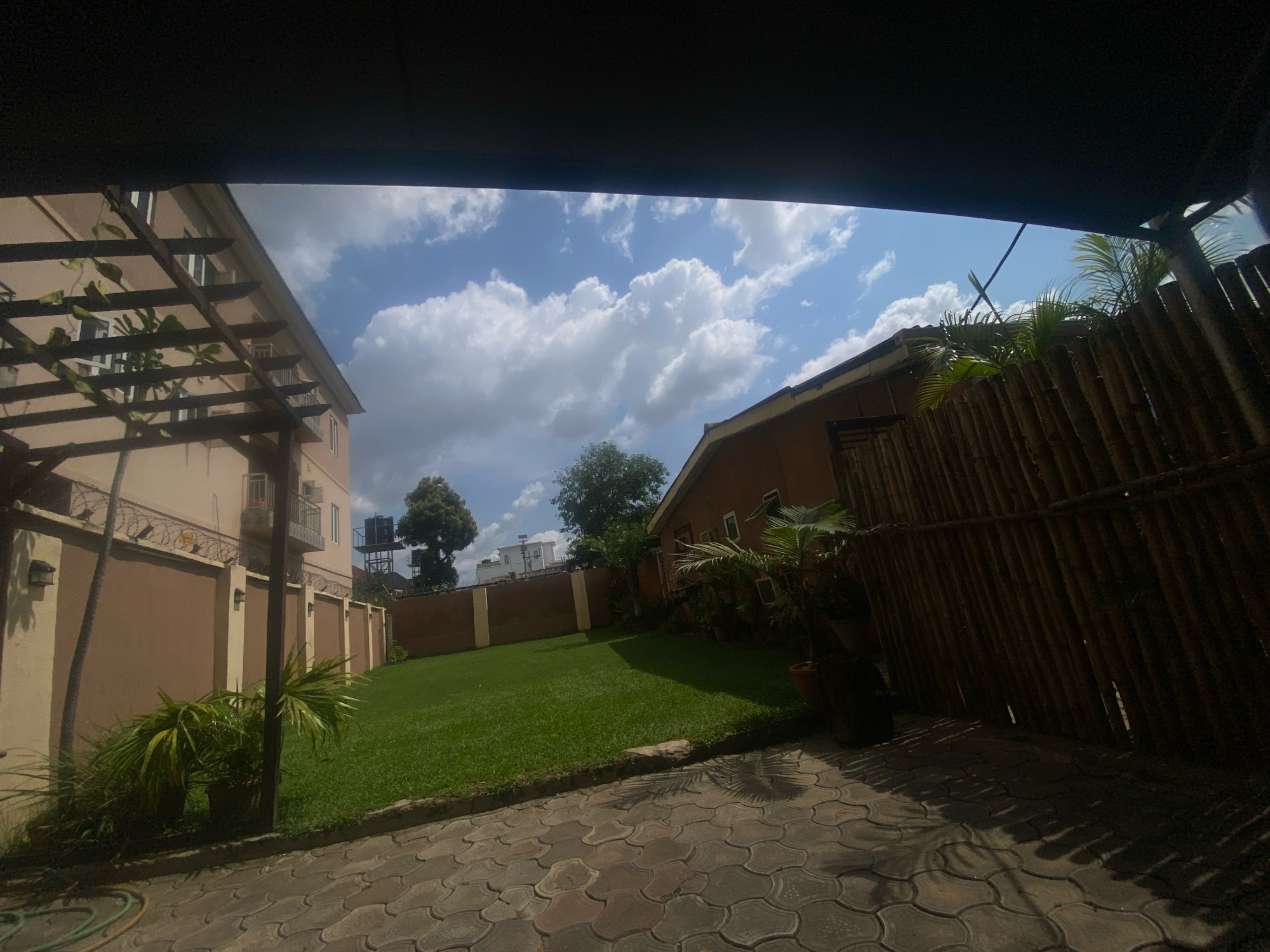 The image captures a bright, sunny day in a residential courtyard, likely located in Barden Yana, Nigeria. In the foreground, a paved area with interlocking concrete stones leads into a well-maintained green lawn. To the left, a multi-story building with a beige facade is partially visible, featuring a wooden pergola structure extending over a section of the yard. This building has balconies and windows, with air conditioning units mounted on the exterior. A brown wall, topped with barbed wire, encloses the property.
To the right, a fence constructed from vertical bamboo poles separates the courtyard from an adjacent area. Several potted plants, including palm-like foliage, are placed in the courtyard. In the background, another building with an orange-brown exterior is situated, and further back, trees and other structures are visible, suggesting an urban or suburban setting. The sky is blue with scattered, fluffy white clouds. The scene appears to be empty of people.