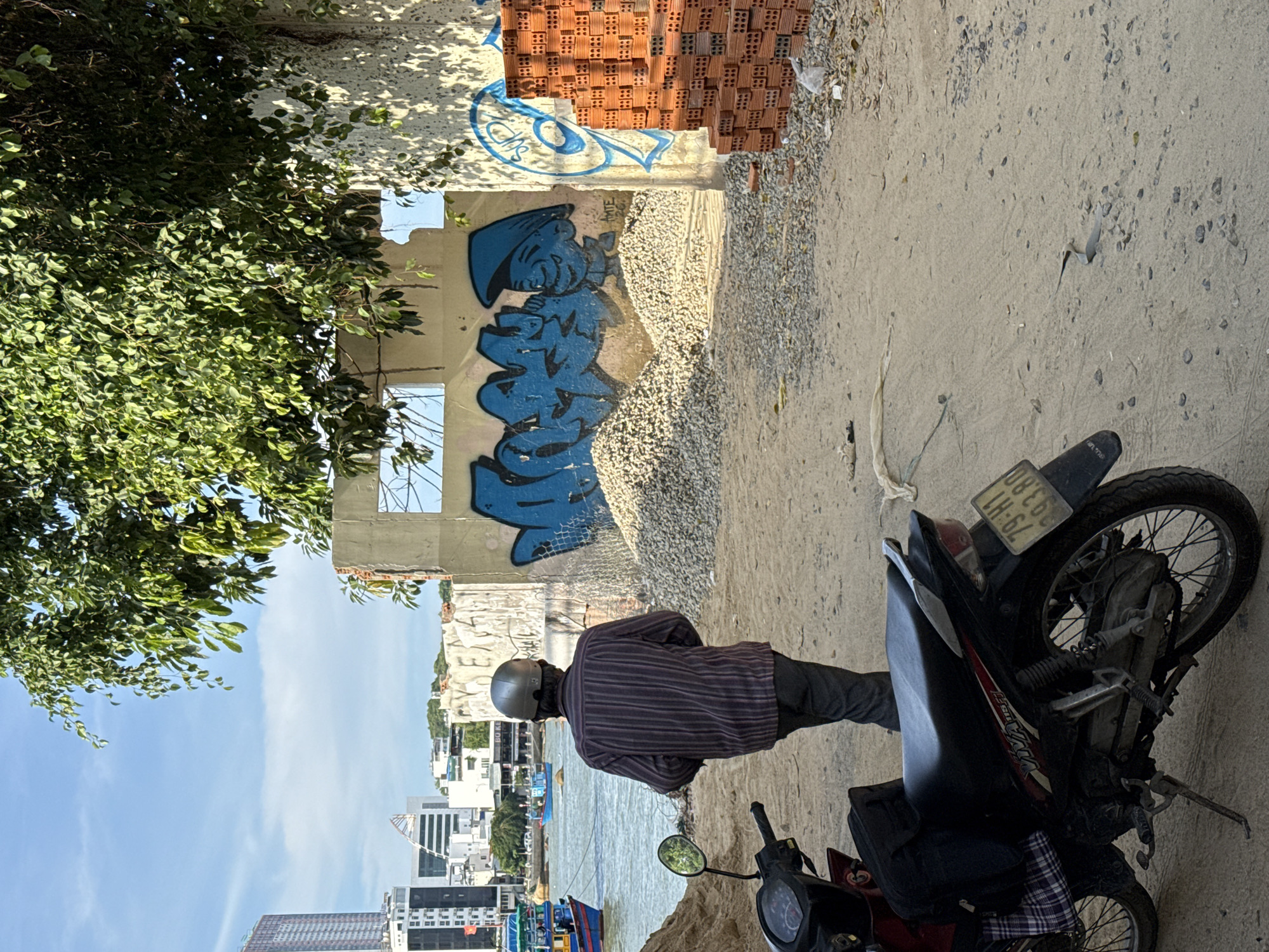 The image depicts an outdoor scene in Bac Nha Trang Ward, Vietnam. In the foreground, a red and black motorcycle is parked on a dirt path. A man wearing a striped shirt, dark pants, and a silver helmet is walking away from the camera, towards a body of water. Behind the man, a partially constructed concrete wall features blue graffiti and a stack of red bricks. A pile of gravel is situated in front of the wall. In the background, several buildings and a body of water with boats are visible. The sky is blue with some clouds.