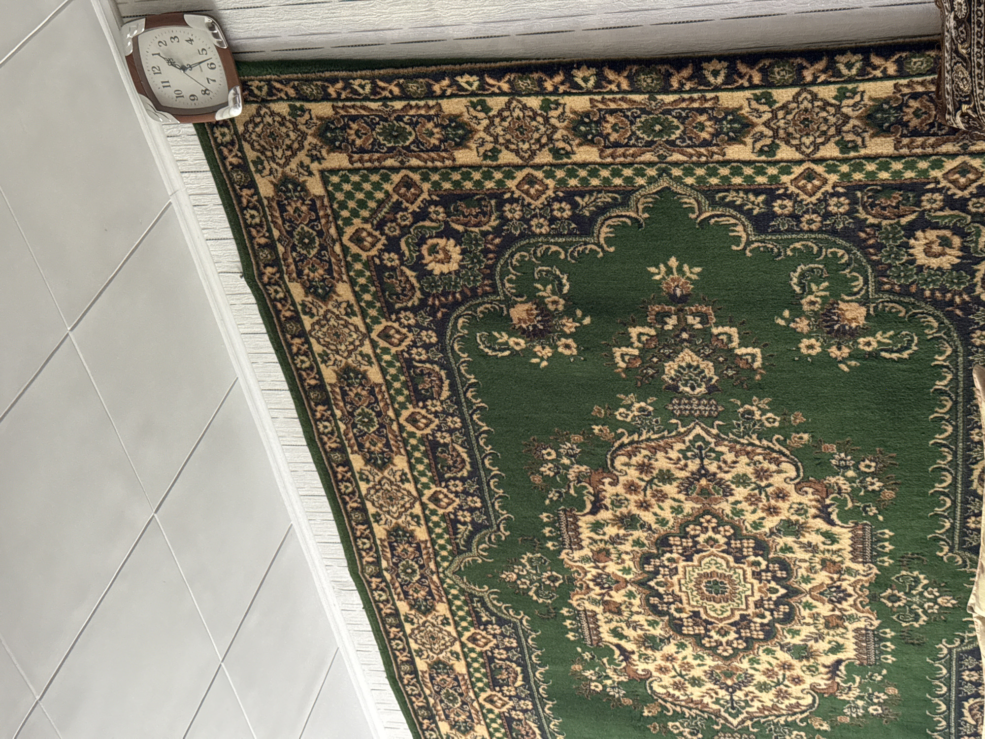 The image shows a section of a room with a large, ornate green rug hanging on the wall. The rug has a complex pattern in shades of gold, blue, and brown. Above the rug is a white, paneled ceiling and a white baseboard. To the right of the rug, a square clock with a wood-tone frame is mounted on a wall covered with vertical white paneling. The clock has a white face with black numbers and hands. The environment appears to be indoors, possibly a residential space, given the decorative rug and wall clock. No people or specific landmarks related to Shymkent, Kazakhstan, are visible in the image.