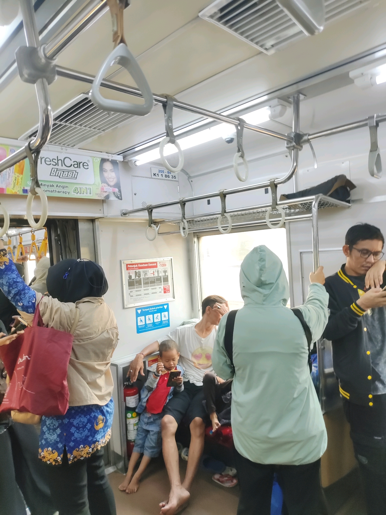 The image depicts the interior of a crowded commuter train, likely in East Jakarta, Indonesia, as indicated by a sign reading "Tempat duduk prioritas" (priority seating) with Indonesian text and cultural clothing patterns visible on a woman in the foreground. Multiple individuals are standing and seated within the train car, holding onto overhead handles and poles for stability. A child is seated on an adult's lap, engrossed in a mobile phone. Another man in glasses is looking at his phone. The train car features fluorescent lighting, ventilation grates, and safety signage. A fire extinguisher is visible near the seated passengers. The overall scene conveys the typical daily commute experience in a densely populated urban environment.