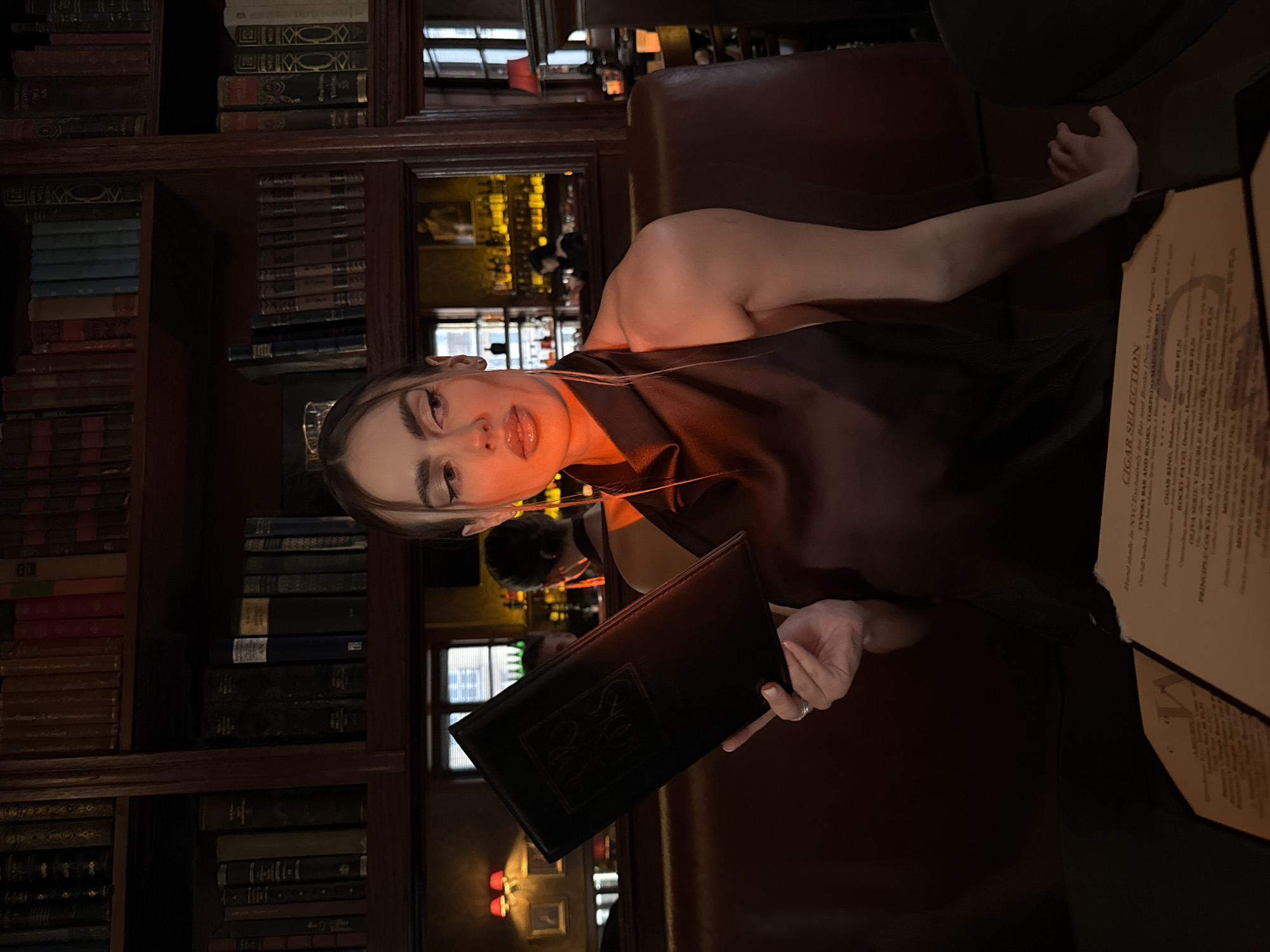 Here is a description of the image:
A woman is sitting at a table in what appears to be a bar or lounge setting, possibly a cigar bar, as suggested by the menu titled "Cigar Selection". The setting features dark wood bookcases filled with books, creating a classic and sophisticated ambiance. The woman holds a black menu with an emblem on it. She is facing the camera. The menu on the table has text that indicates cigar offerings. The environment suggests an upscale establishment.