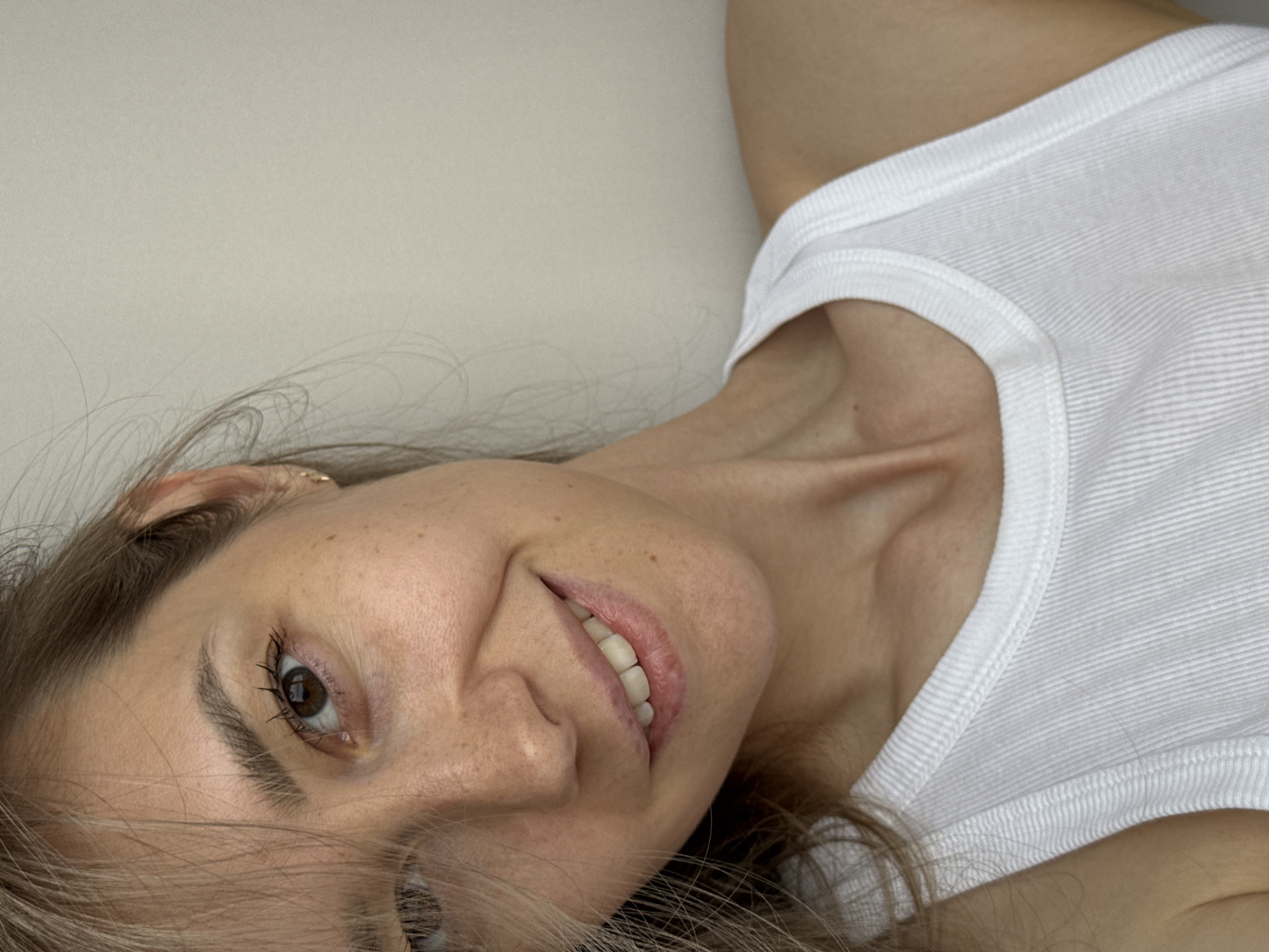 A woman with light brown hair and fair skin is lying on a surface, wearing a white ribbed tank top. She is smiling slightly and looking directly at the camera. Her right eye is clearly visible and brown. The background is a neutral, off-white color, suggesting a wall or ceiling. There is no evidence to suggest the image was taken in the city of Chaikovsky, Russia, or any other specific location.