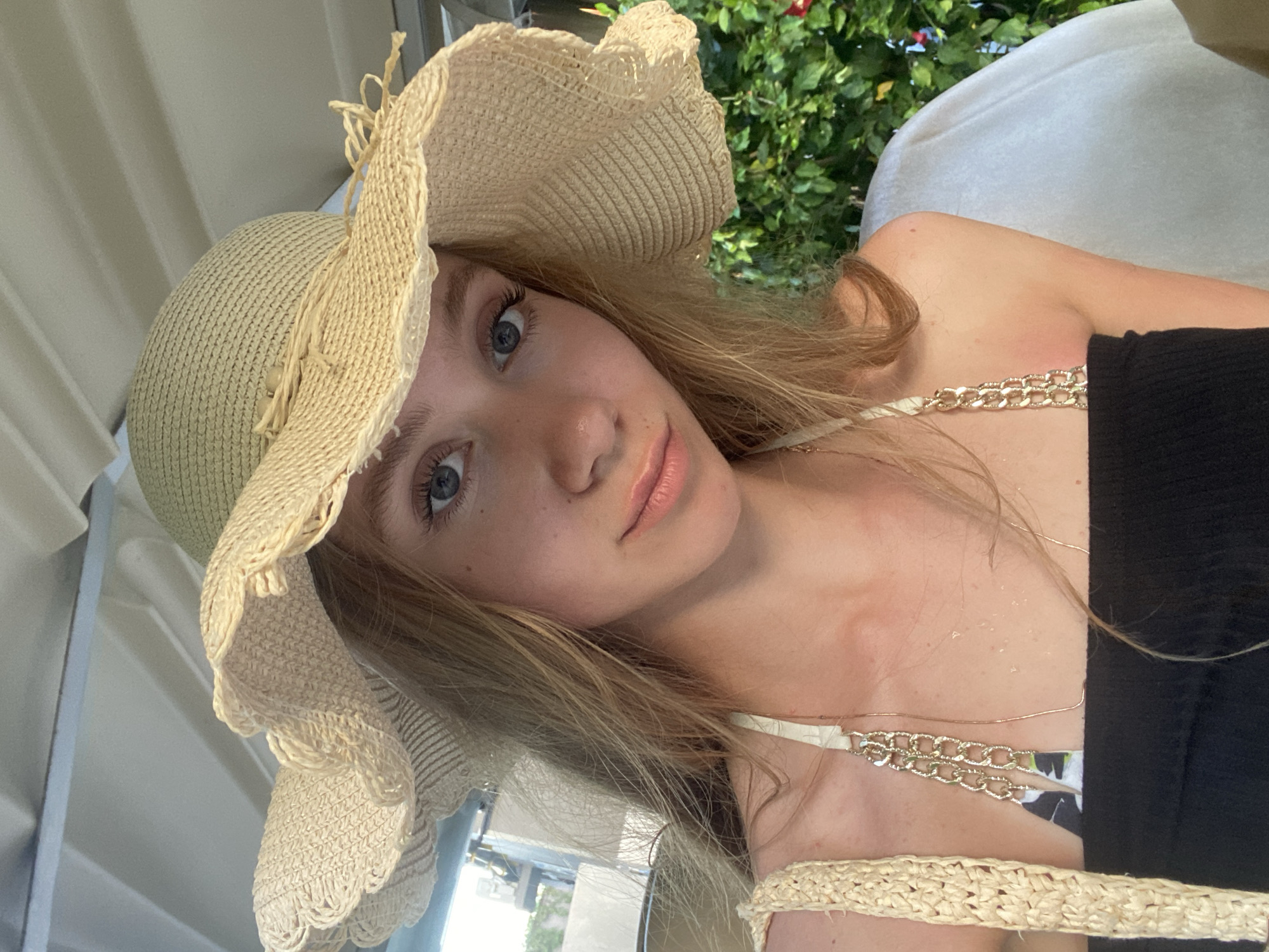 A young woman with fair skin, light brown hair, and blue eyes is pictured in a selfie. She is wearing a wide-brimmed straw hat and a black top with a gold chain strap. She appears to be outdoors, with green foliage visible in the background. The location is Kuta Selatan, Indonesia.
