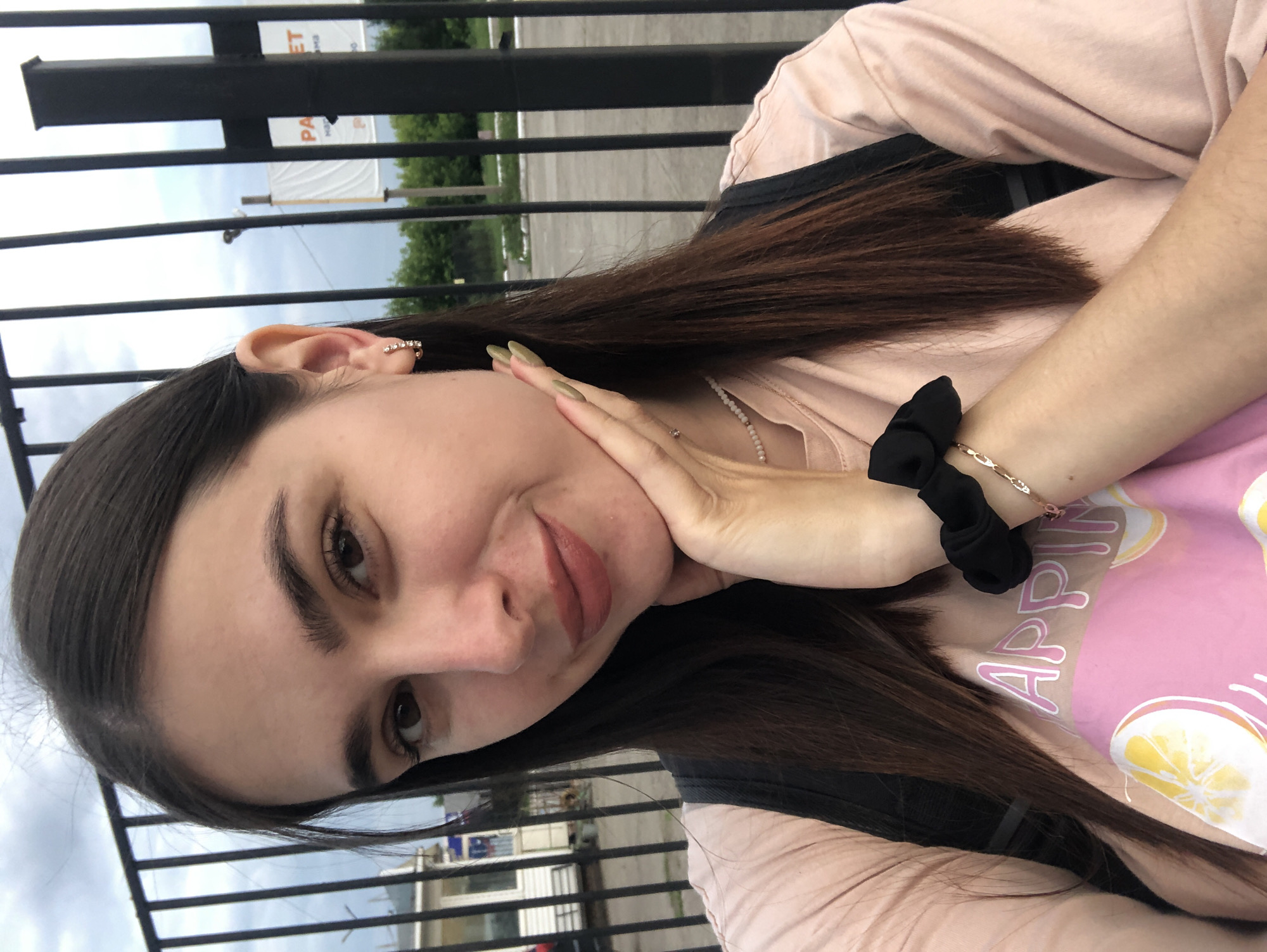 The image is a close-up, low-angle, outdoor selfie of a young woman. She has long, dark brown hair, brown eyes, and is wearing light pink lipstick. Her right hand is resting on her chin, and she is wearing a light pink t-shirt with a graphic print on it. A black scrunchie is visible on her wrist, and she is also wearing a delicate beaded necklace and a small hoop earring with rhinestones in her left ear. Behind her, a black metal fence is visible, with trees and a cloudy sky in the background. A partially visible white banner with text in Russian is also present behind the fence, suggesting a public or commercial area. The overall lighting suggests it is daytime. There are no elements in the image that specifically indicate the city of Chaikovsky, Russia.