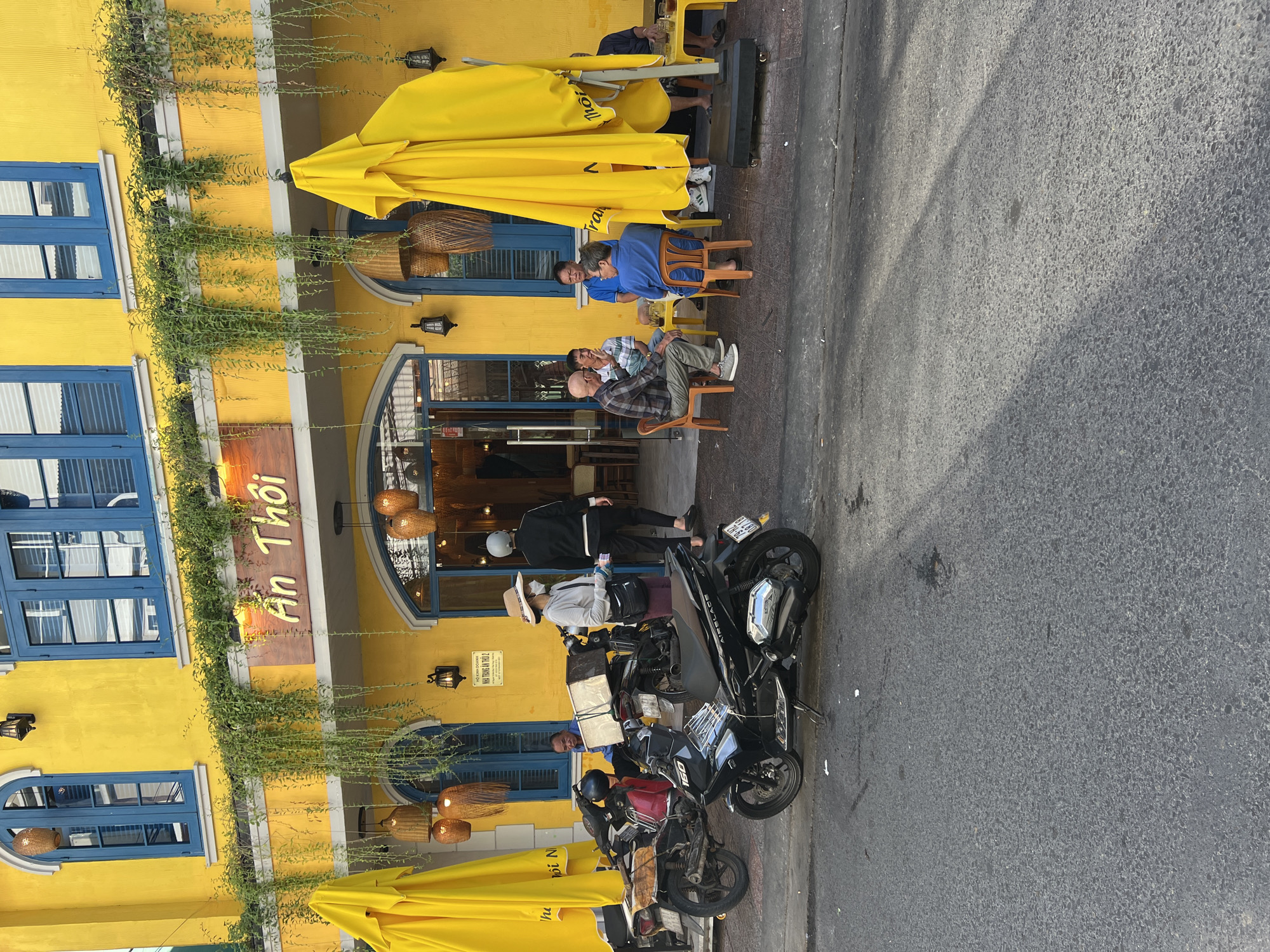 The image depicts a street-level view of a brightly colored yellow building with blue-trimmed windows, characteristic of architecture found in cities like Nha Trang, Vietnam. Green vines cascade down from the building's facade. A sign above the entrance reads "An Thoi" in a stylized font.
In the foreground, a black scooter is parked on the asphalt street. To the left of the scooter, a red scooter is also parked. Further down the street, several people are seated at small tables outside the building, engaged in conversation and consuming drinks. One person is wearing a hat and a face mask, and another is wearing a helmet. The overall scene suggests a casual outdoor cafe or restaurant setting on a sunny day, with shadows indicating late afternoon light. The presence of multiple scooters is typical for urban street scenes in Vietnam.
