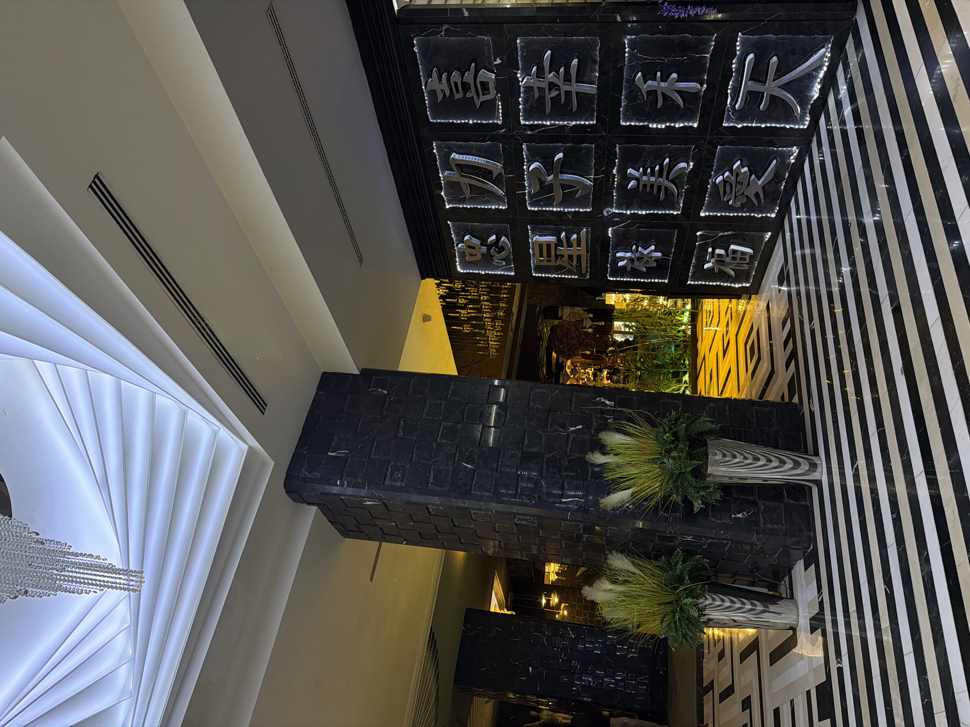 The image shows the interior of a building with Asian decor. The ceiling has illuminated Chinese characters on black square tiles. A rectangular column covered in small, dark tiles stands below the characters. Two large decorative planters with tall grass sit on top of the column. The floor is patterned with black and white stripes. A modern, layered, white lighting fixture hangs from another part of the ceiling. The location in the city of Sofia, Bulgaria, cannot be definitively determined from the visual evidence.